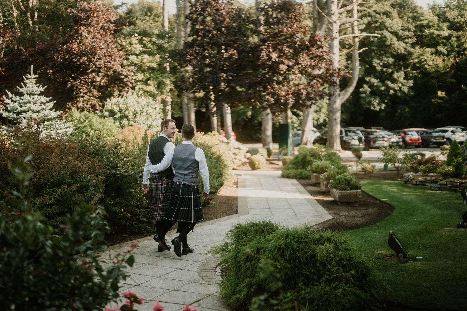 Two people in kilts walk along a garden path surrounded by lush landscaping.