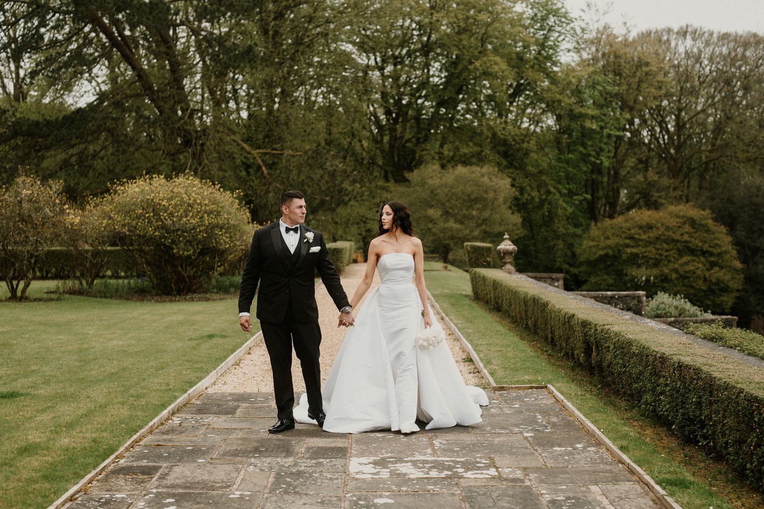 A couple walks hand in hand along a stone pathway in formal wedding attire.