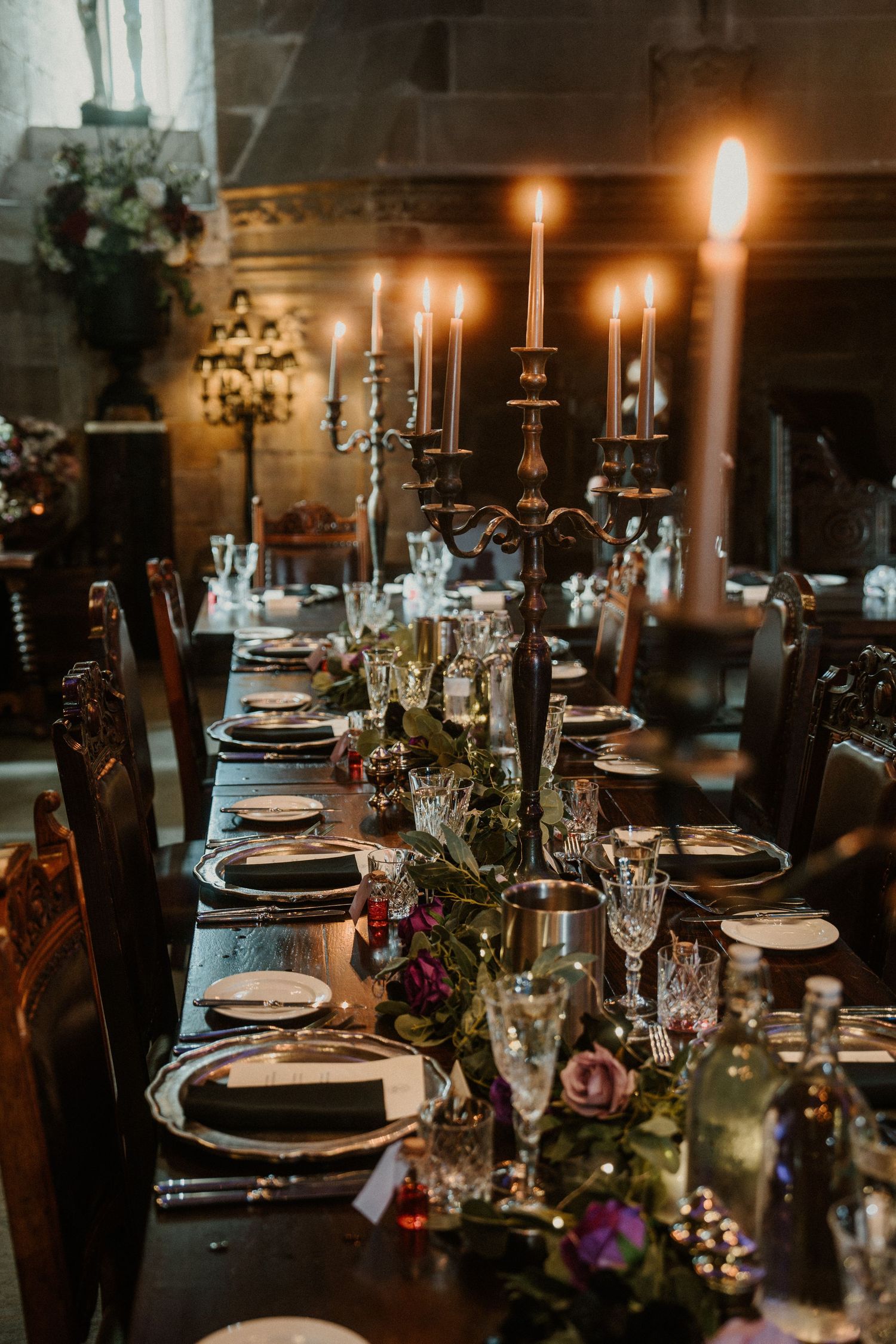 Elegant candlelit dining table with dark place settings, crystal glasses and romantic floral centerpieces at dusk.