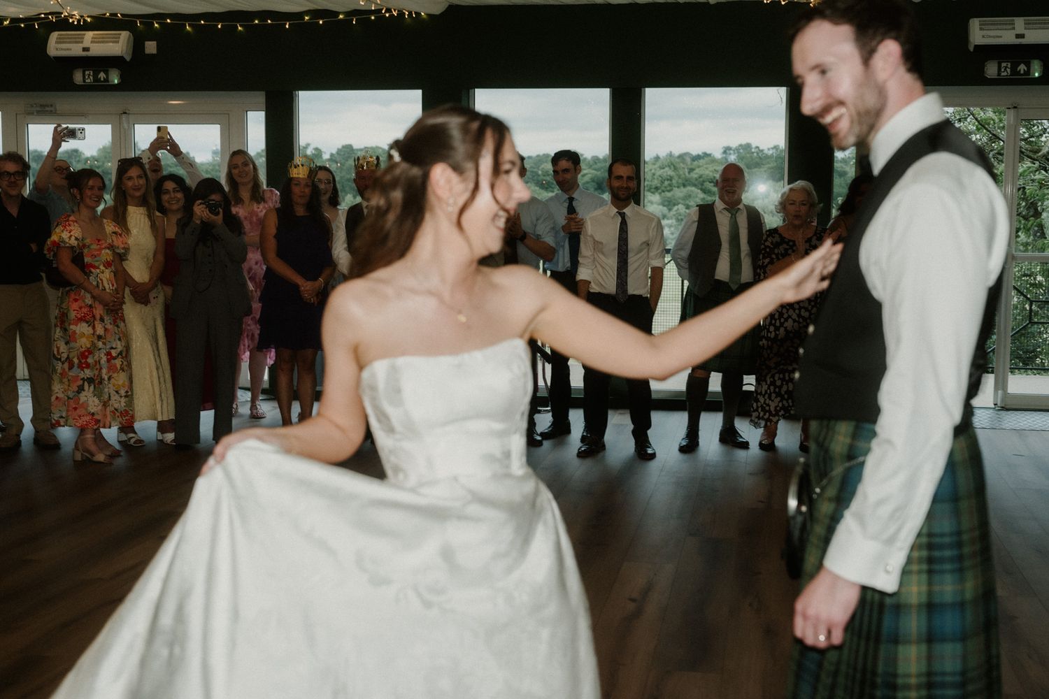 Bride in white strapless gown dances with groom in tartan kilt during first dance at wedding reception.