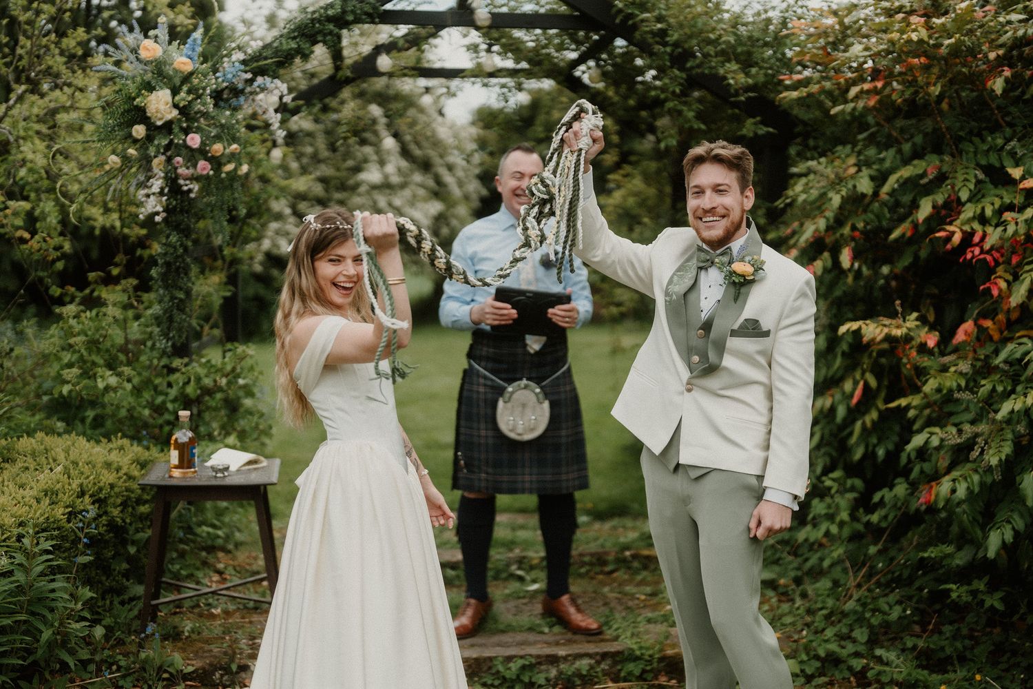 Joyful newlyweds celebrate handfasting ceremony outdoors under floral arch with kilted officiant in lush garden setting.