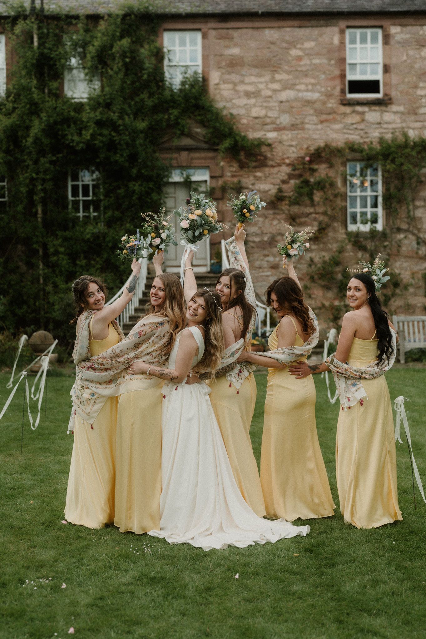 Bride and bridesmaids in yellow gowns celebrate outdoors, raising bouquets joyfully in front of a stone manor.