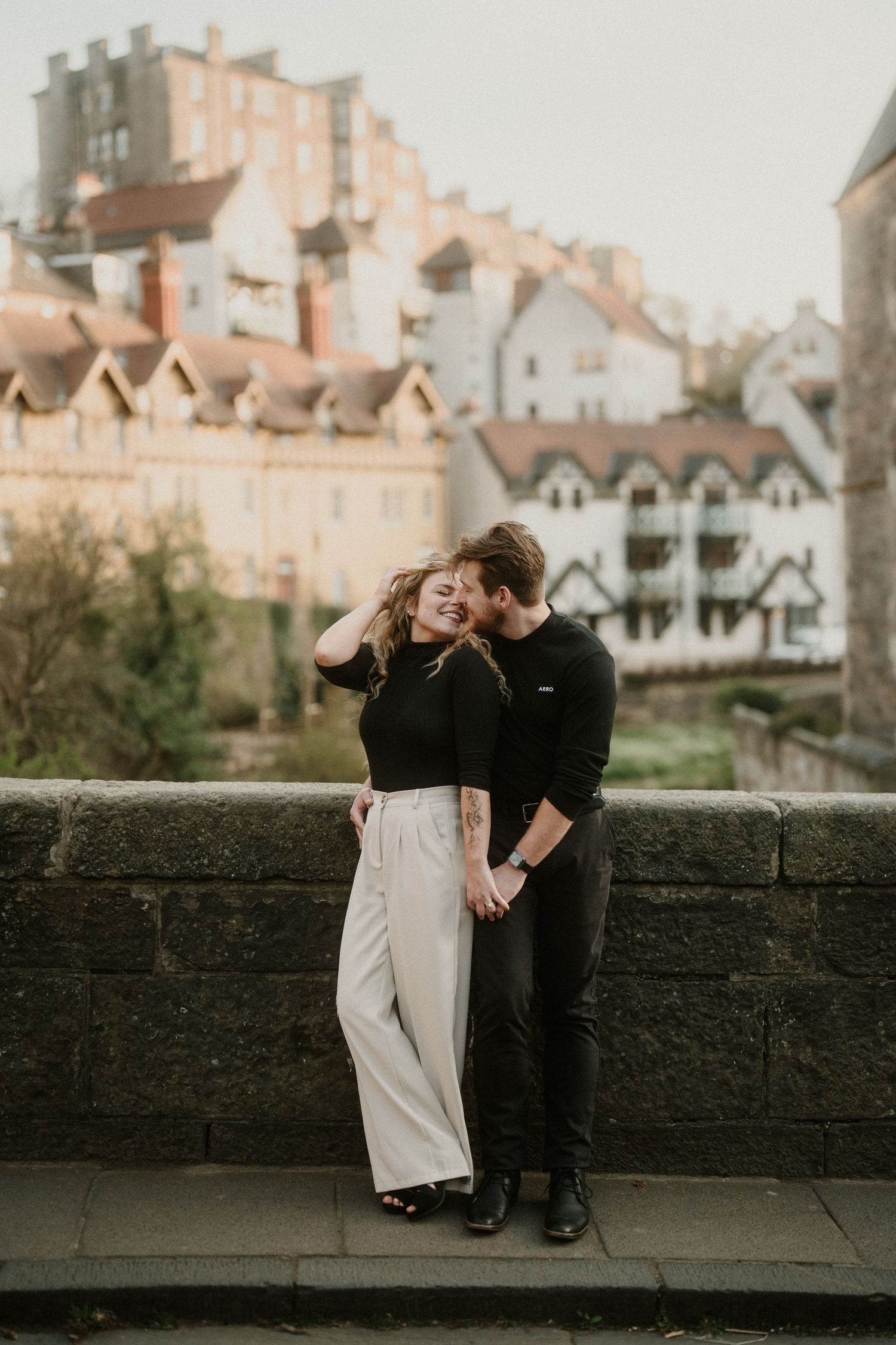 A couple embraces romantically against a stone wall with historic buildings in the background during golden hour.