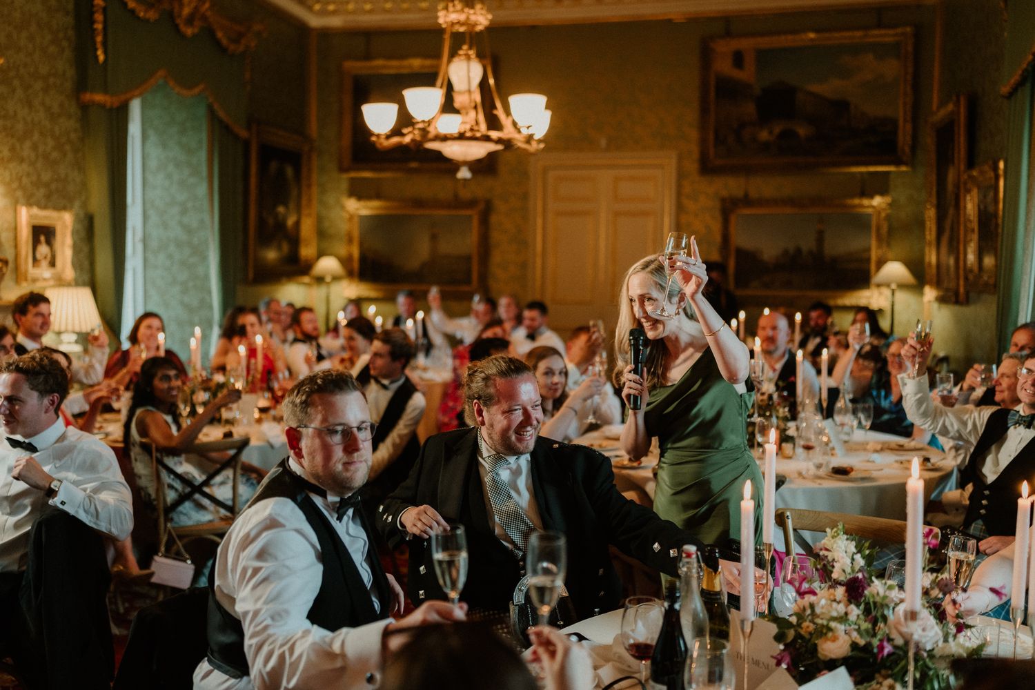 Elegant wedding reception in a grand dining room with chandelier, guests in formal attire raising glasses in celebration.