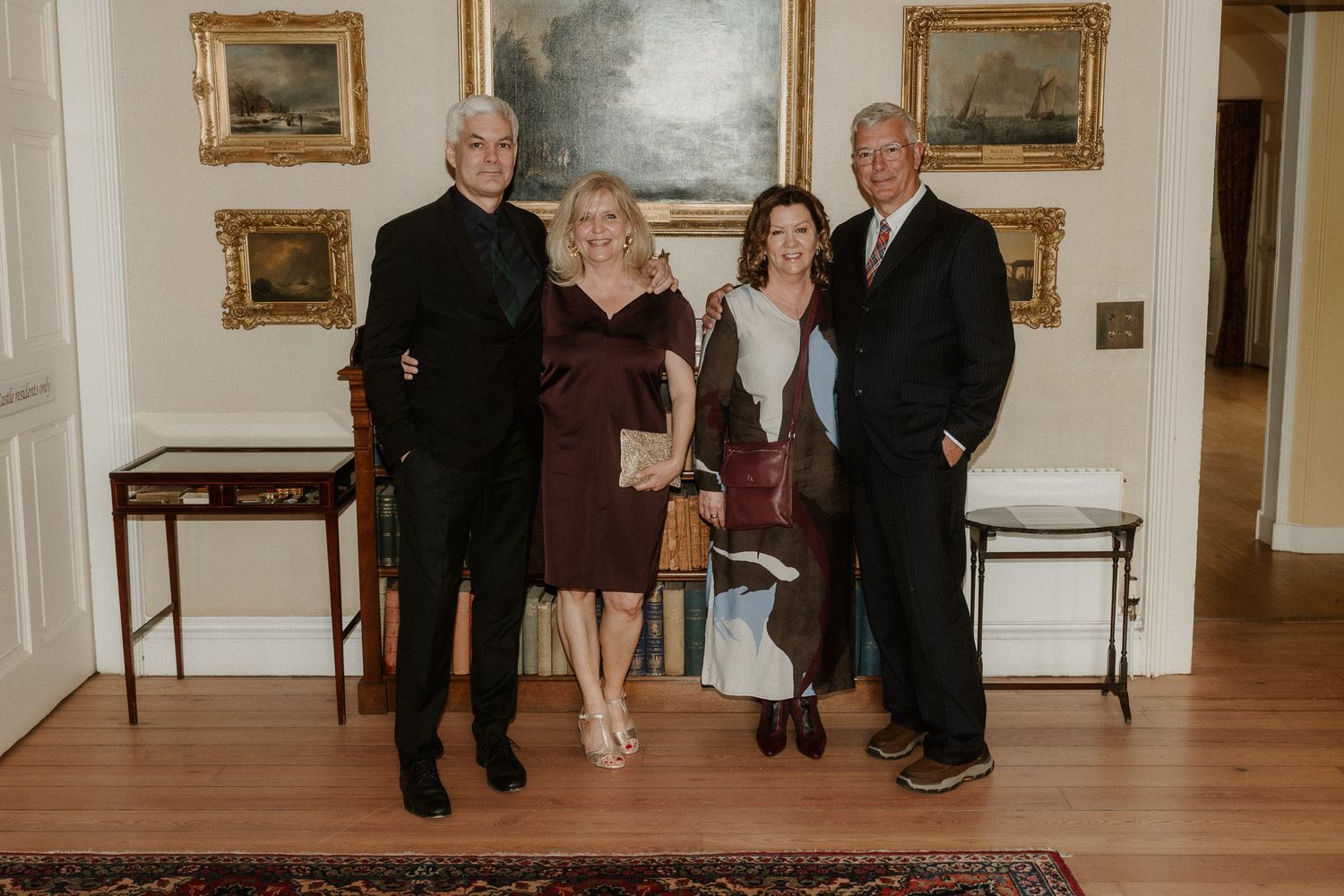 Four elegantly dressed adults posing together in a classic interior with hardwood floors and framed paintings.