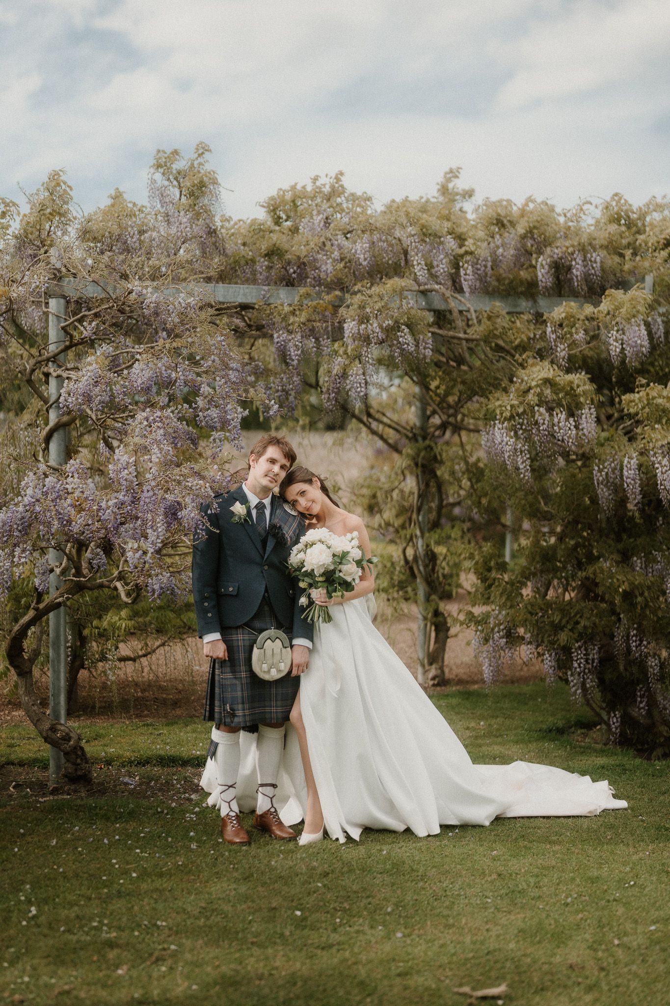 Bride and groom in Scottish kilt pose under purple wisteria pergola on their wedding day.