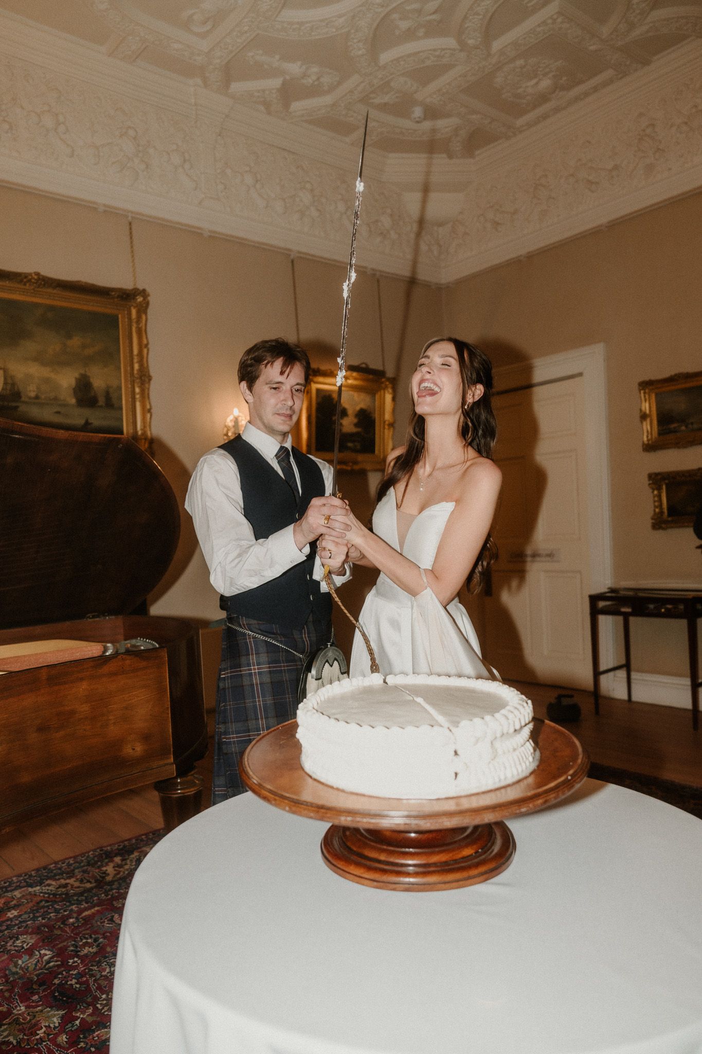 Bride and groom cutting white wedding cake together in elegant room with ornate ceiling, sparkler lit on top.