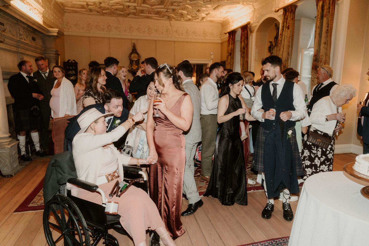 Elegant wedding reception with guests mingling, including a person in a wheelchair, in a grand decorated room.