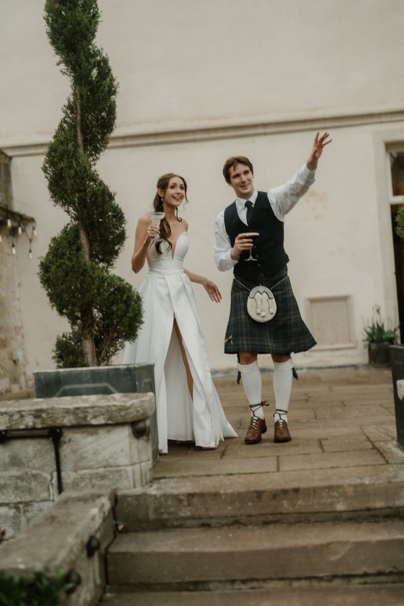 Bride in white gown and groom in Scottish kilt raising glasses on stone steps at elegant wedding venue.
