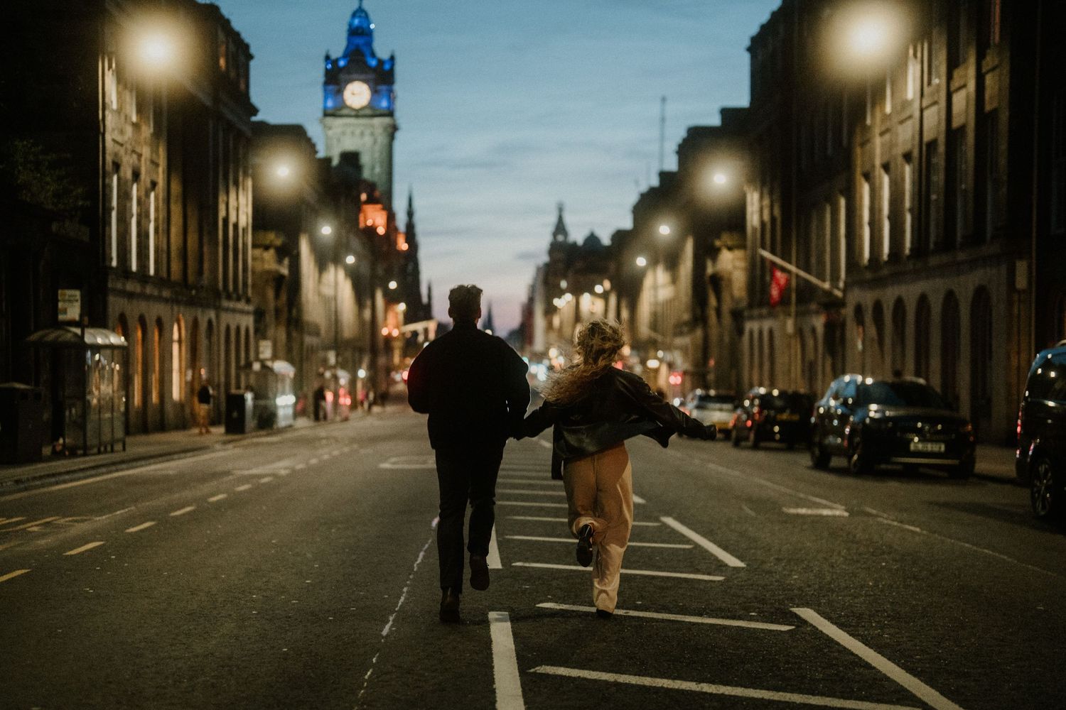 Couple holding hands walking down illuminated city street at night with clock tower.