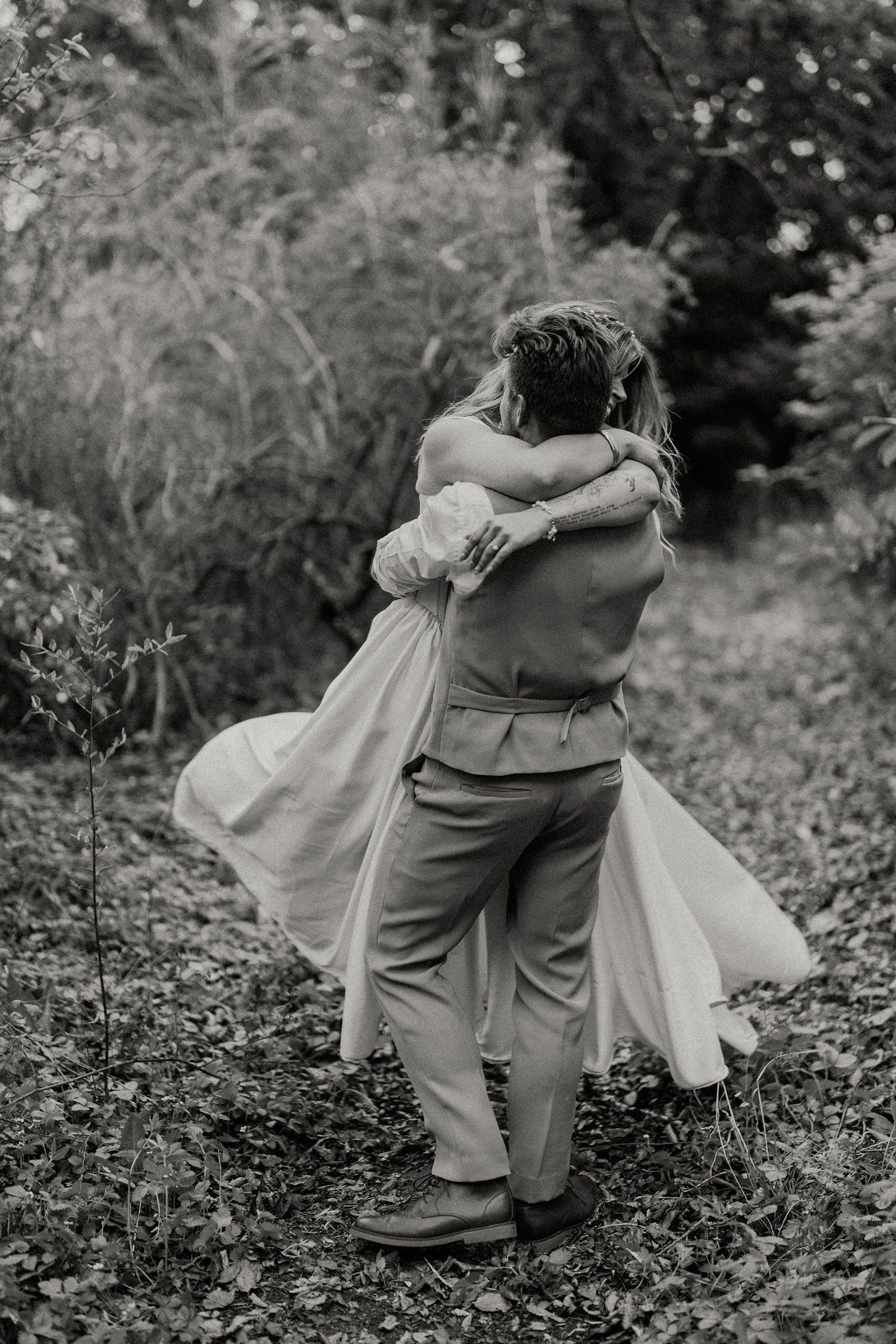 Black and white photo of a couple embracing in a lush garden, bride in flowing dress, groom in suspenders.