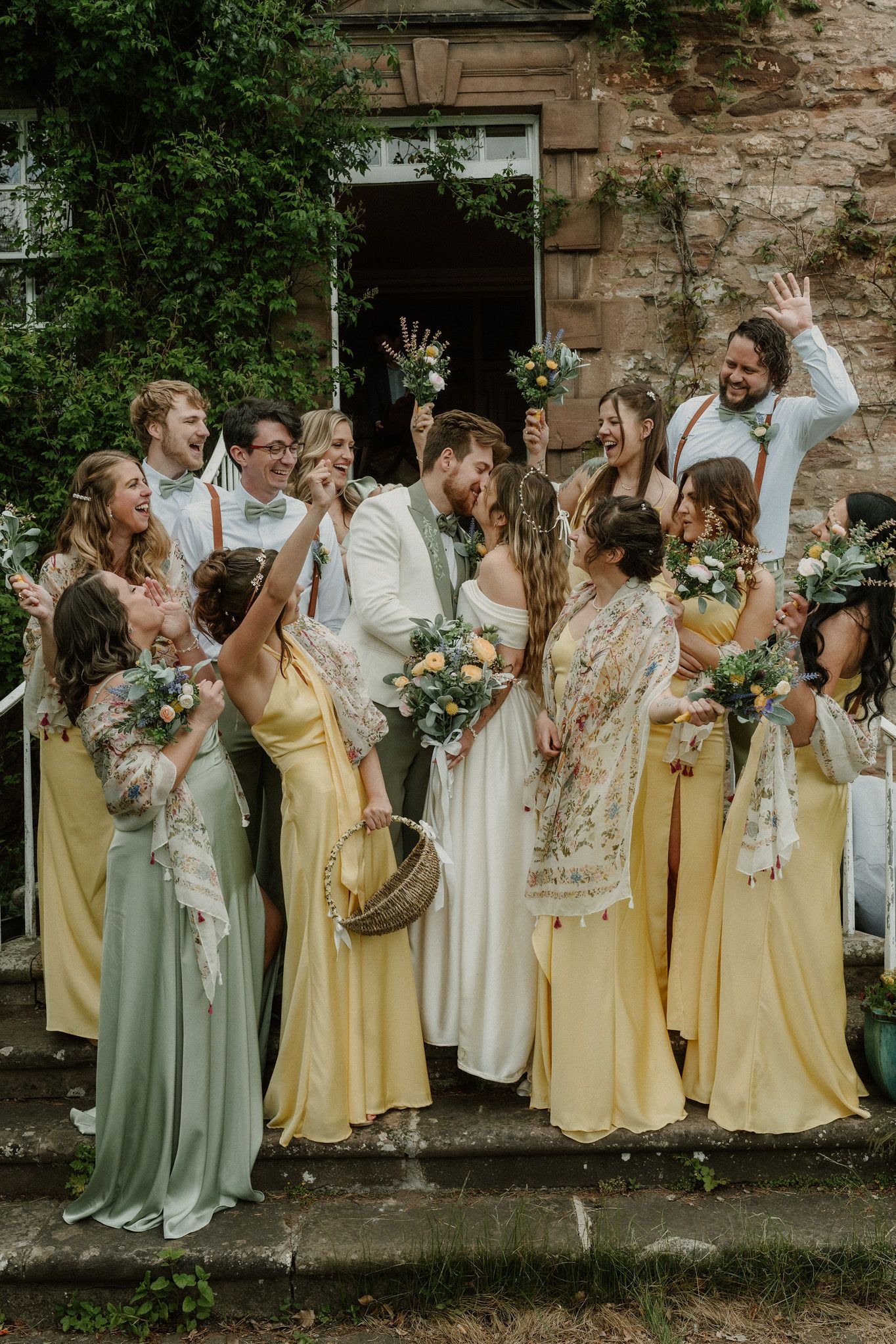 Wedding party celebrating on stone steps, bridesmaids in yellow and sage green gowns with floral bouquets, bride and groom kissing.