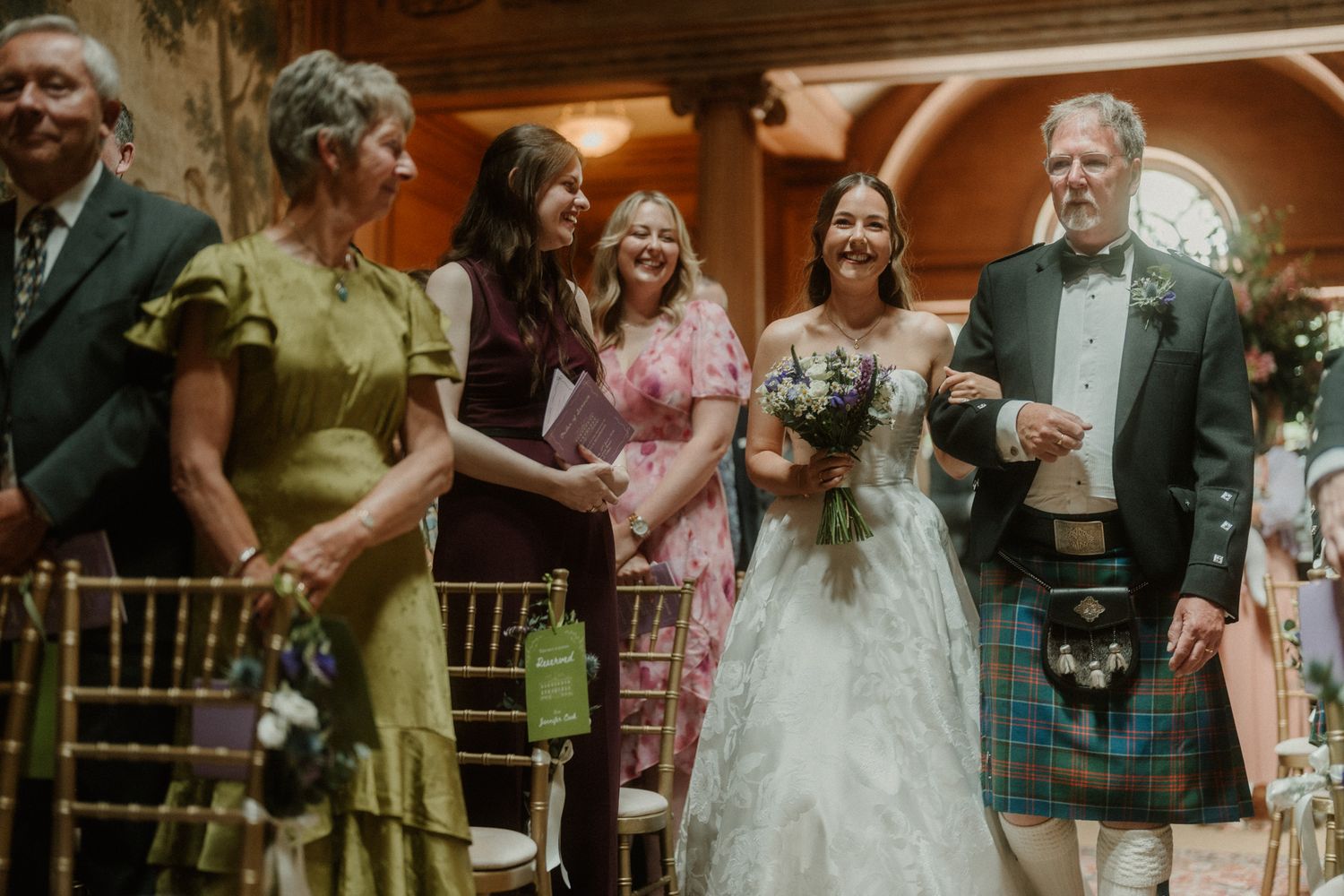 Bride in white gown walks down aisle with man in Scottish kilt, surrounded by smiling guests at elegant wedding ceremony.