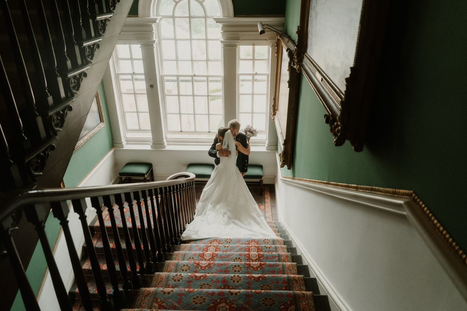 Bride and groom share a romantic moment on a grand staircase with green walls and ornate carpet at a wedding venue.