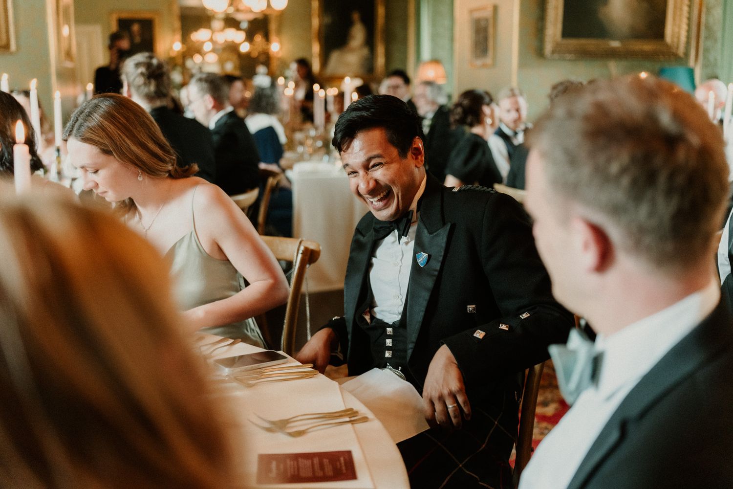 Smiling man in a black dinner jacket and bow tie laughs at a candlelit formal banquet table surrounded by elegantly dressed guests.