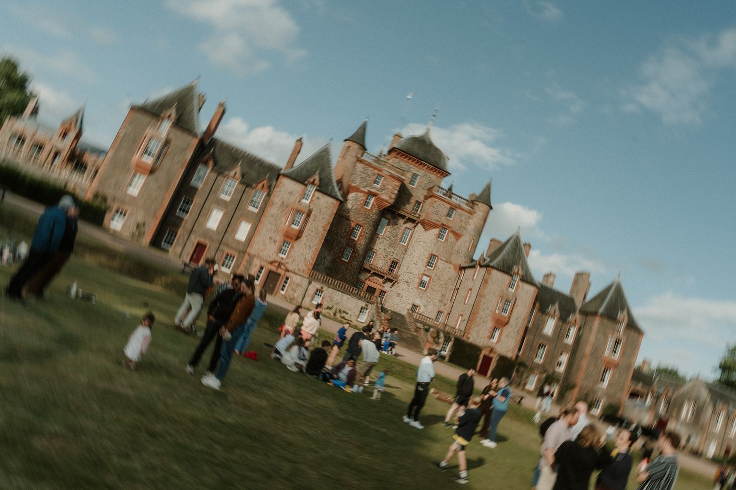 Wedding guests gather on a sunny lawn in front of a grand red brick castle with turrets and steep slate roofs.