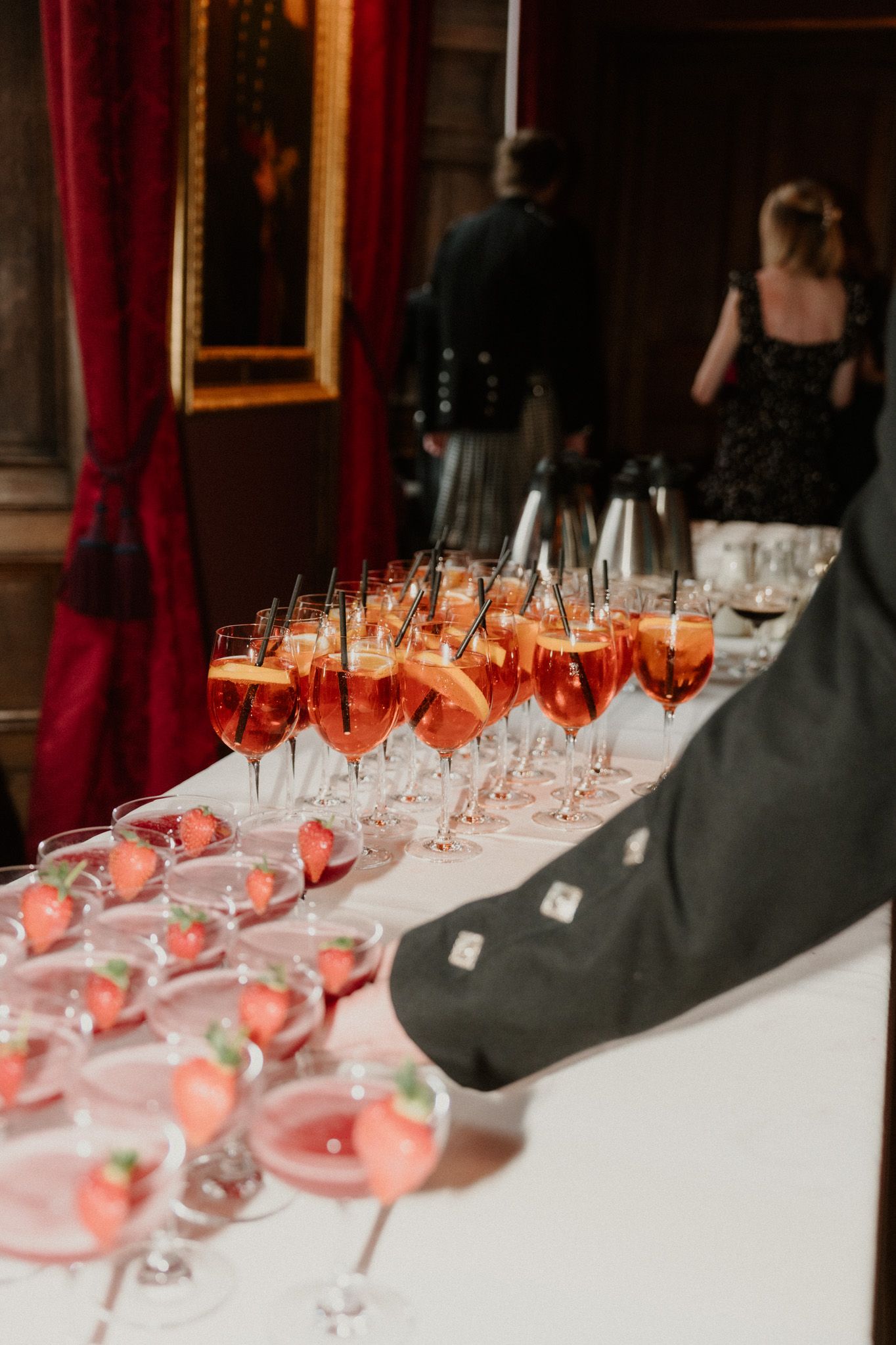 Rows of orange spritz cocktails and strawberry champagne coupes line a white table at an elegant event with red velvet drapes.