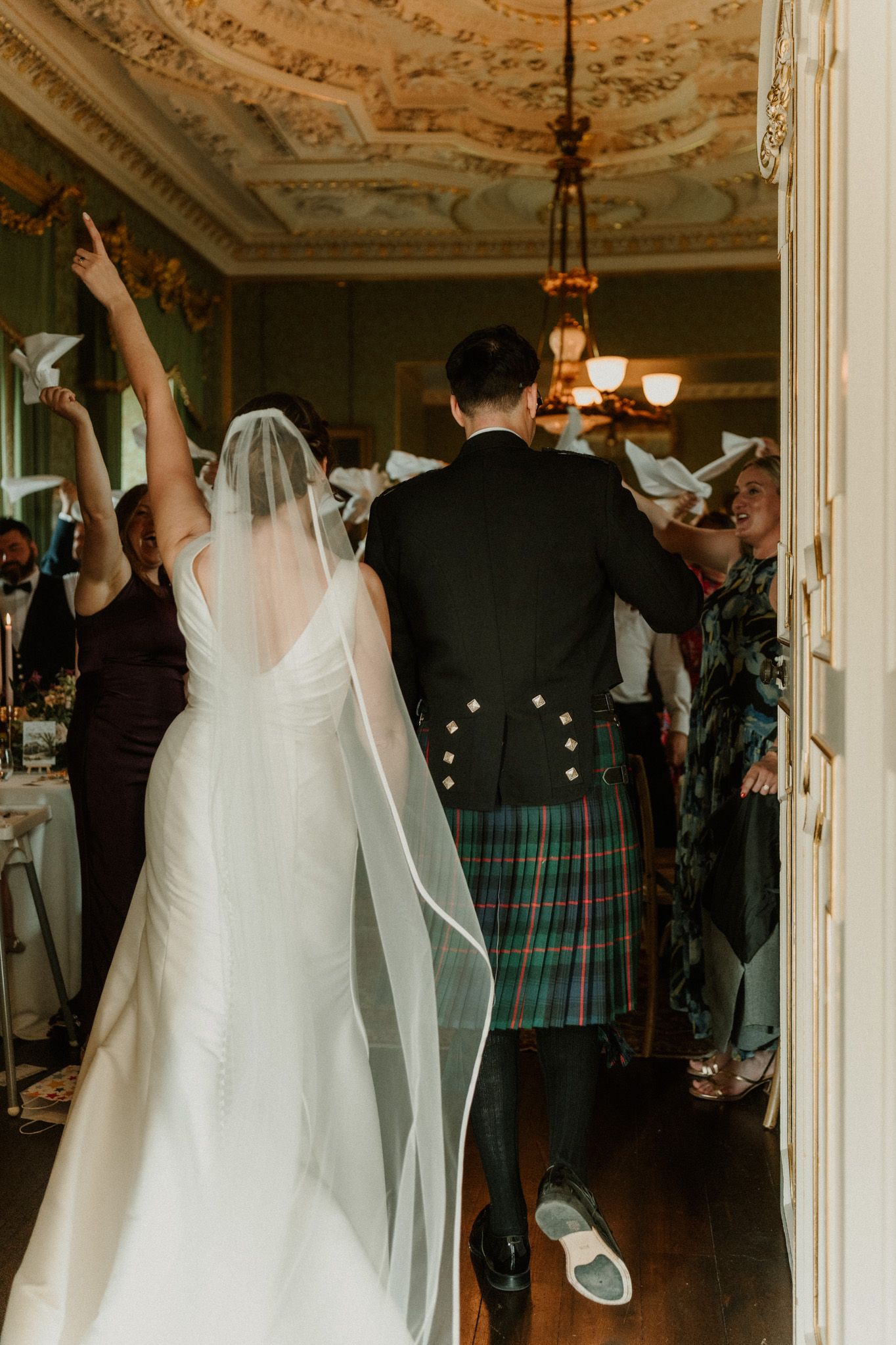 Bride in a long veil and groom in a tartan kilt walk into an ornate reception room as guests cheer and wave white napkins.