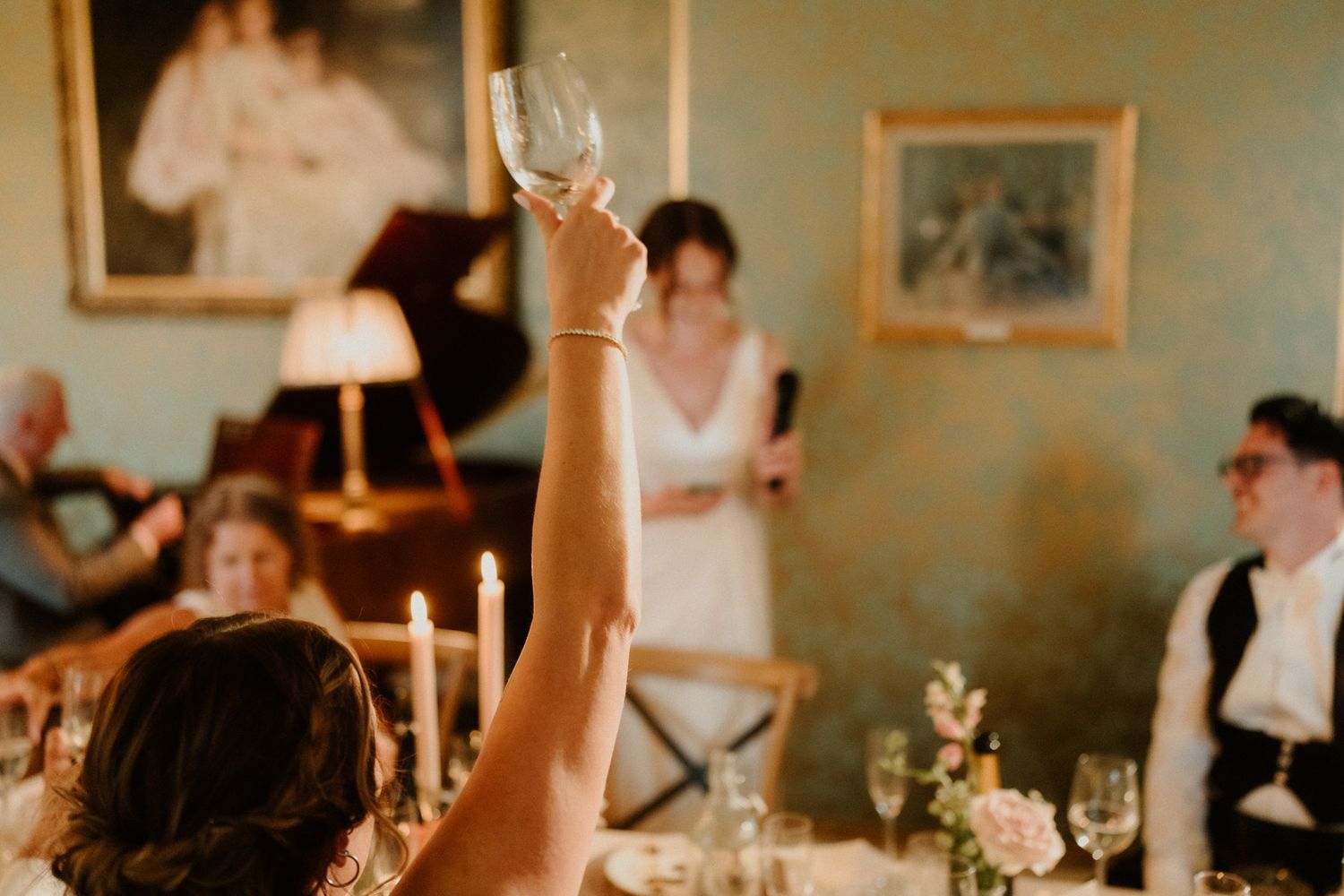 Wedding guest raises a champagne glass in a toast while a woman in white stands near a piano in an elegant dining room with candles.