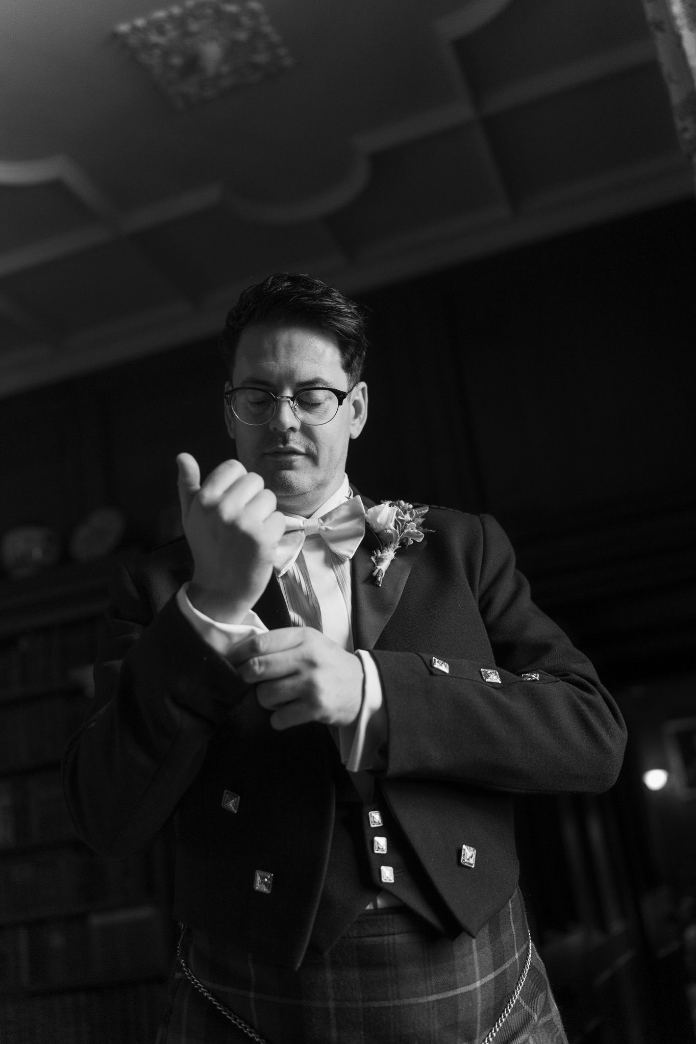 Black and white photo of a groom in glasses, kilt, and bow tie adjusting his shirt cuff, wearing a boutonniere in an elegant, ornate room.