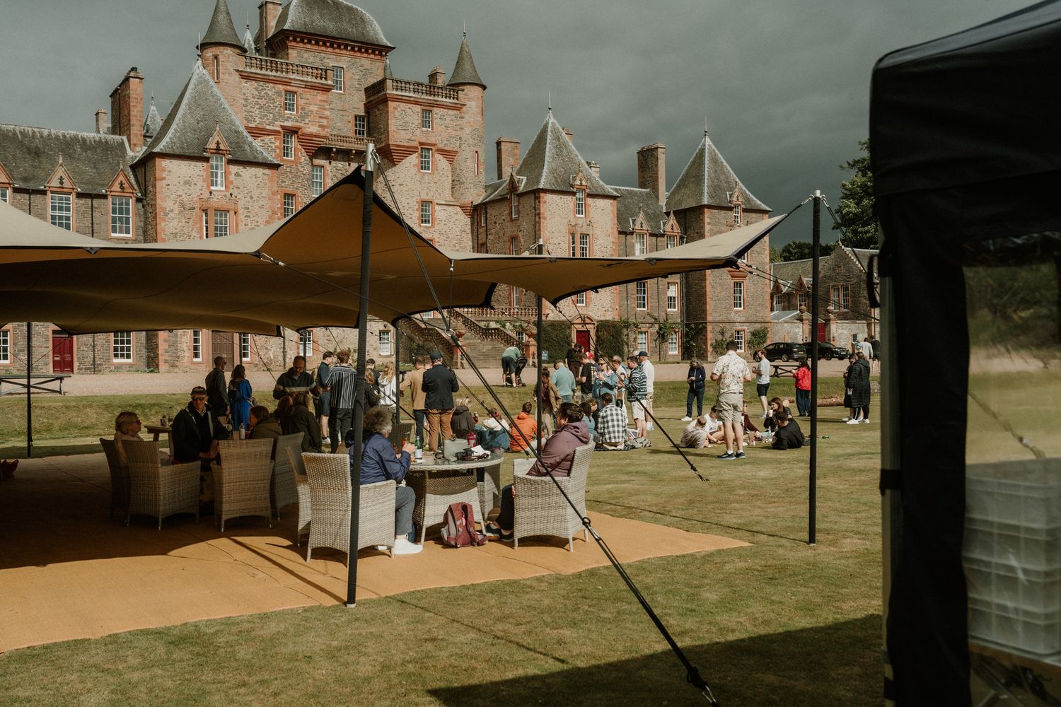 Guests gather under an open tent on a sunny lawn outside a historic stone castle with turrets at an outdoor event.