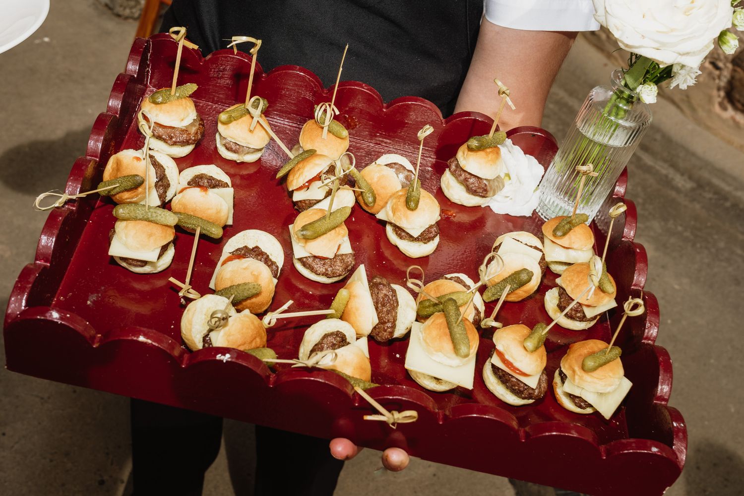 Server holding a red lacquered tray of mini burger sliders topped with pickles and secured with decorative picks, with a flower vase nearby.