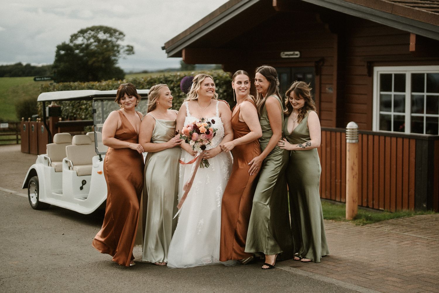 Wedding party in earth-toned dresses poses outside wooden building on overcast day.