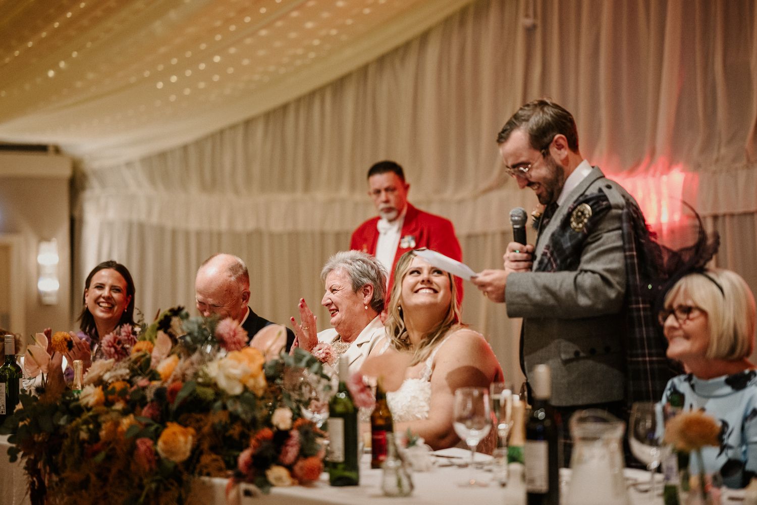 Wedding party enjoying speeches at head table decorated with autumn-colored floral arrangements.