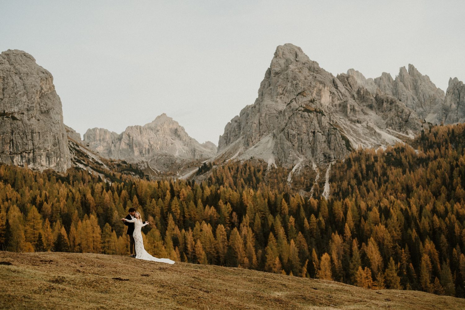 Bride in white dress poses against dramatic mountain landscape with autumn foliage.