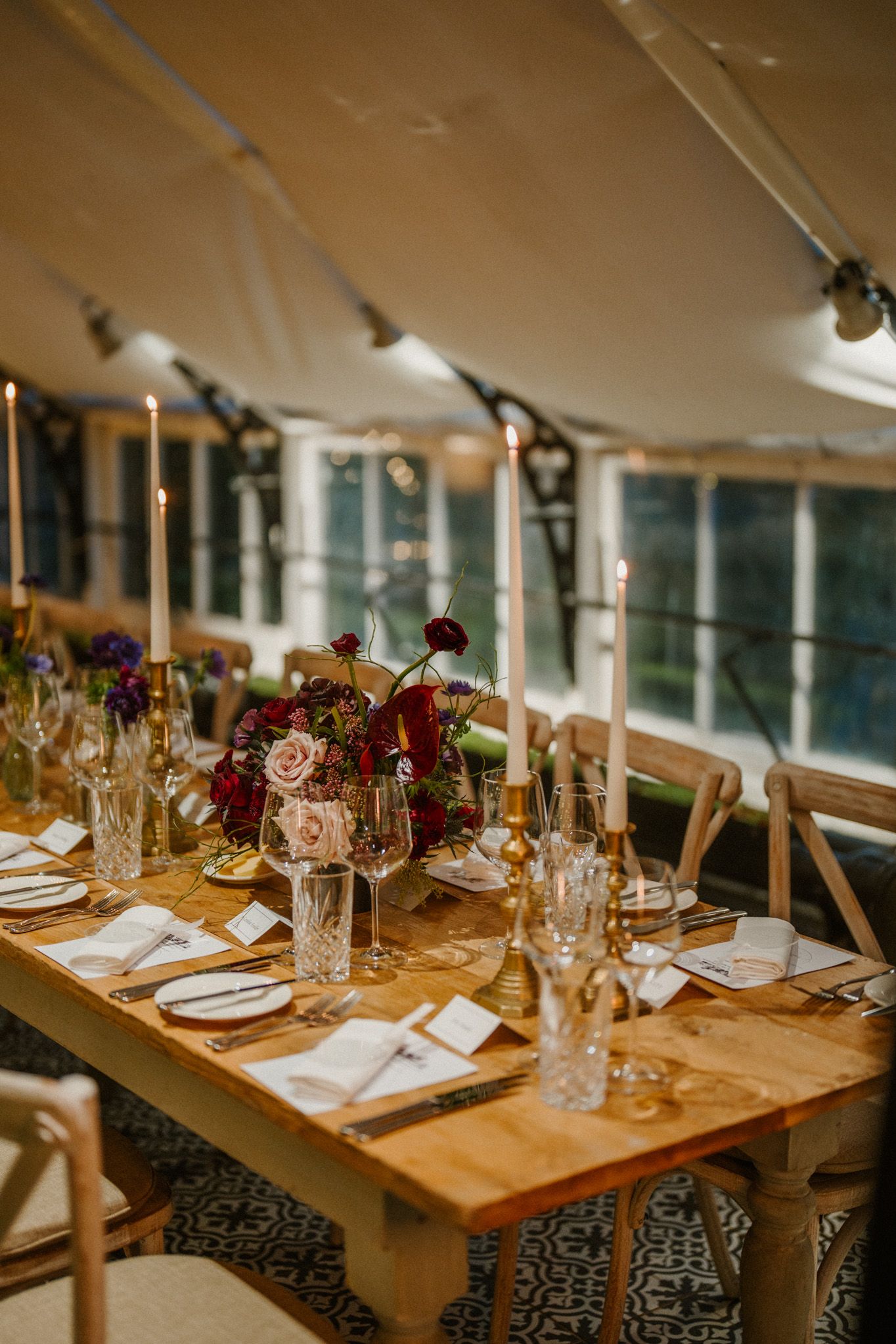 Wedding reception table with burgundy floral centerpiece and crystal glassware under tent lighting.