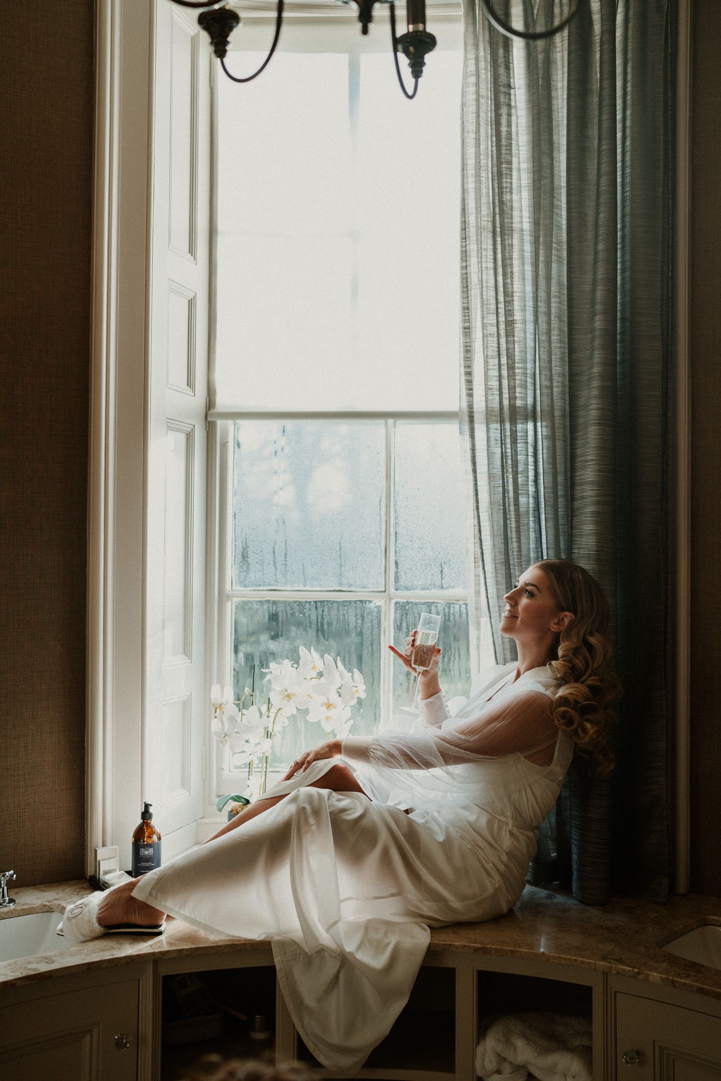 Person in white robe relaxes by window during wedding day preparations.