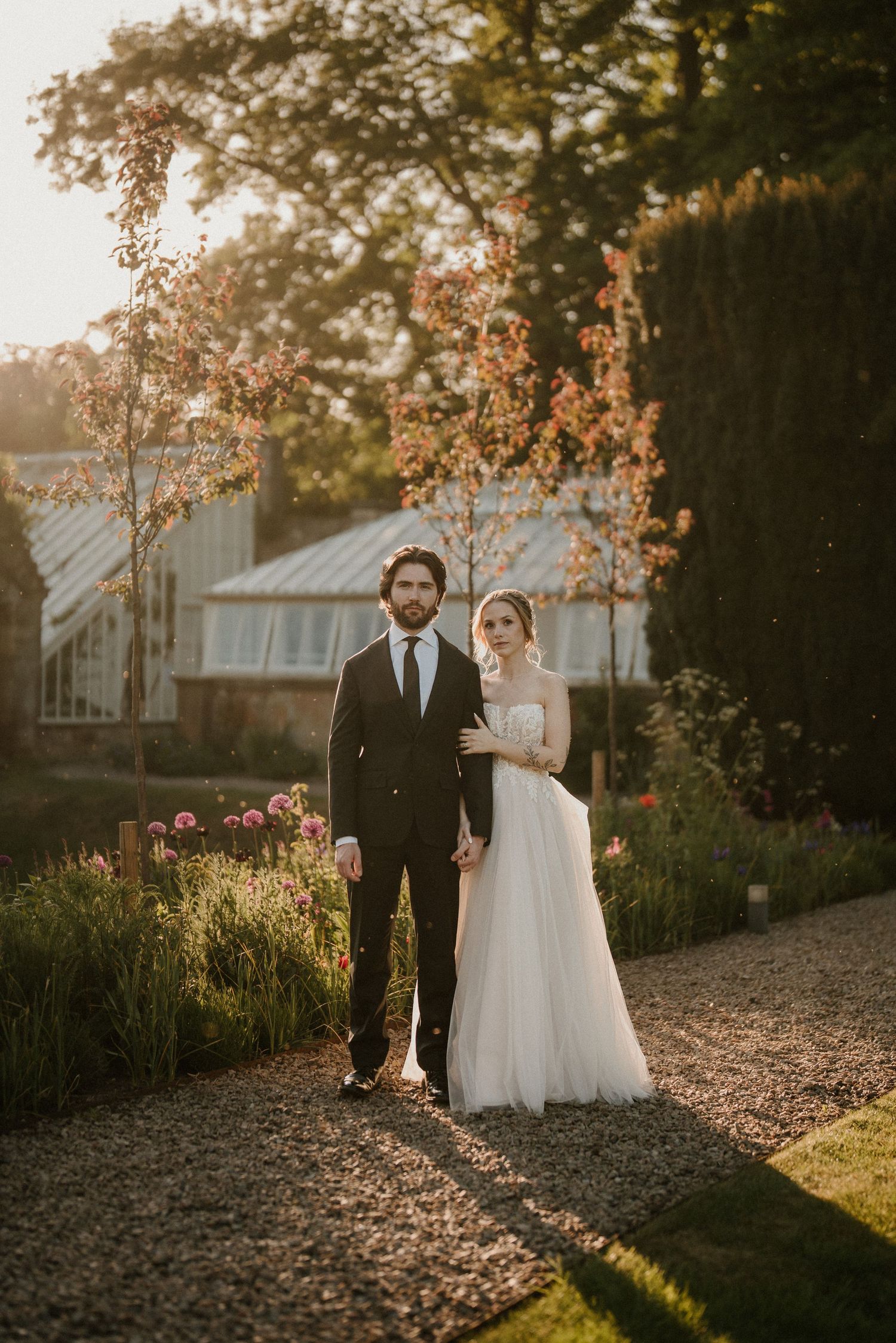 Sunset wedding portrait in garden with greenhouse and golden light.