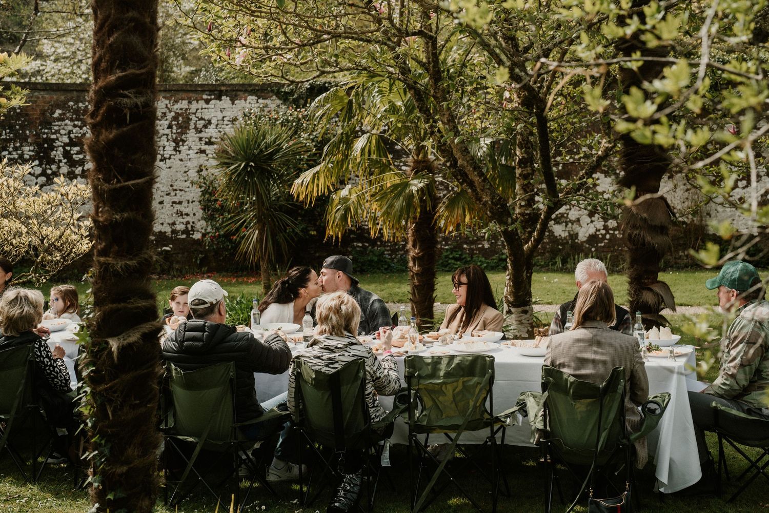 Elegant outdoor dining scene with guests seated at a long white table under palm trees.