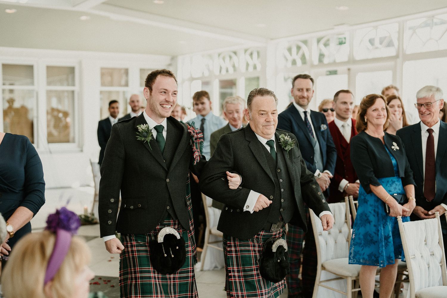 Wedding guests mingle in a bright reception hall with large windows.