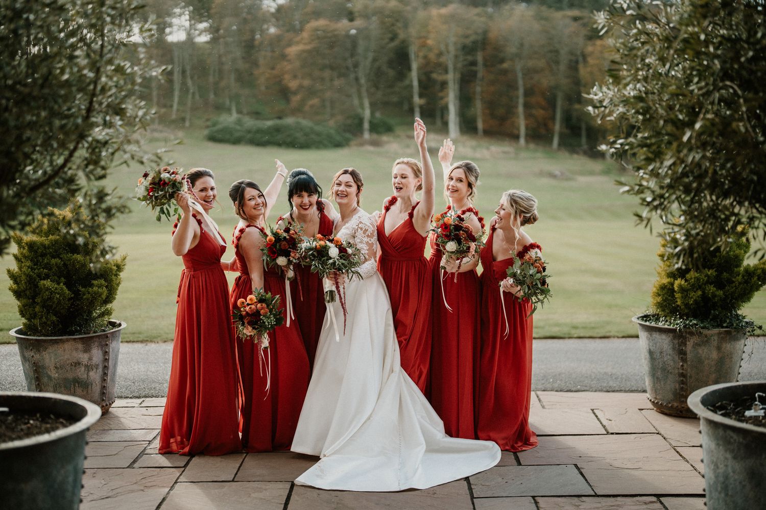 Bridal party in red dresses and white wedding gown stand together outdoors during autumn.