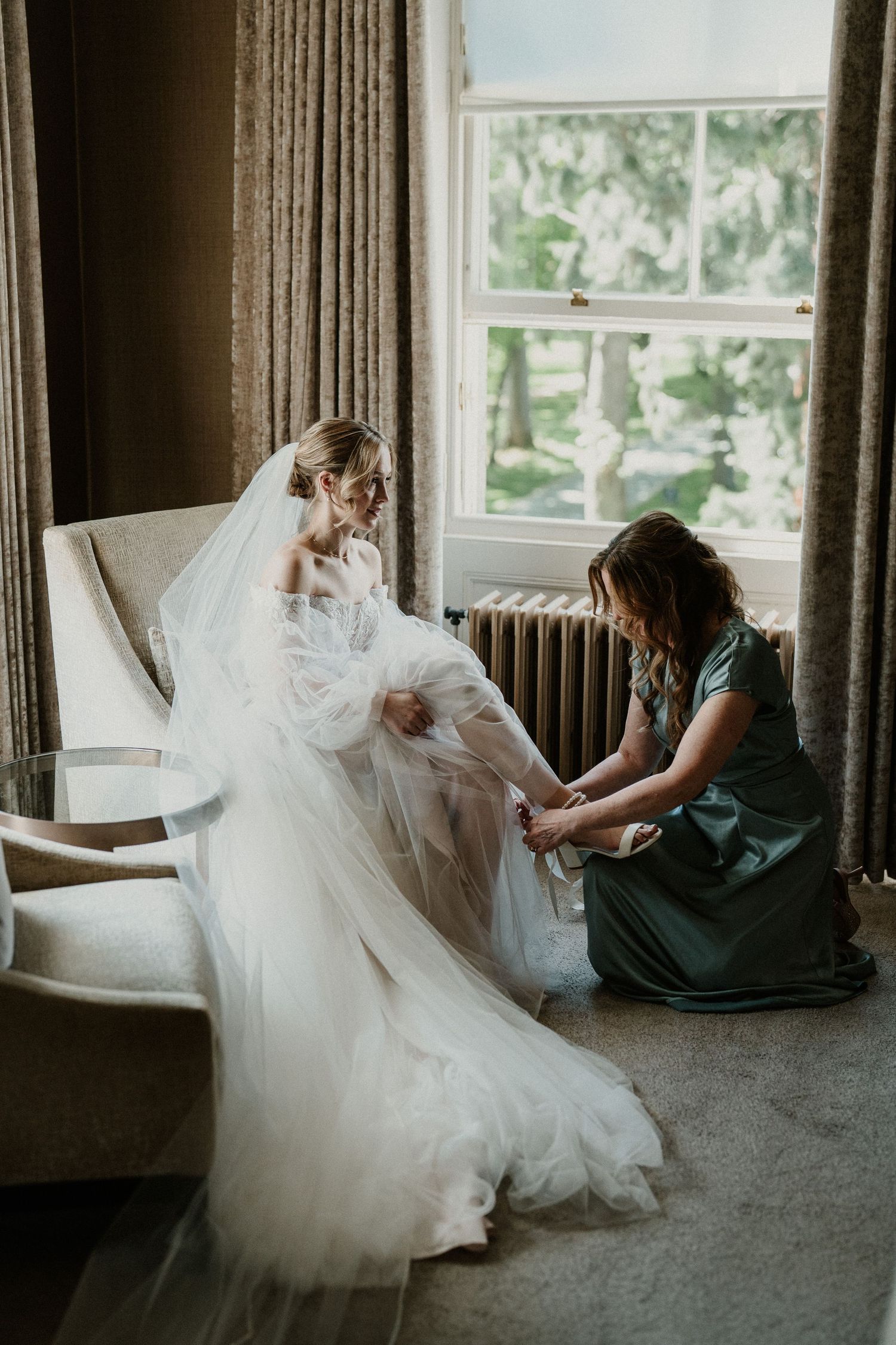 Flowing white wedding dress being arranged on carpeted floor near window.