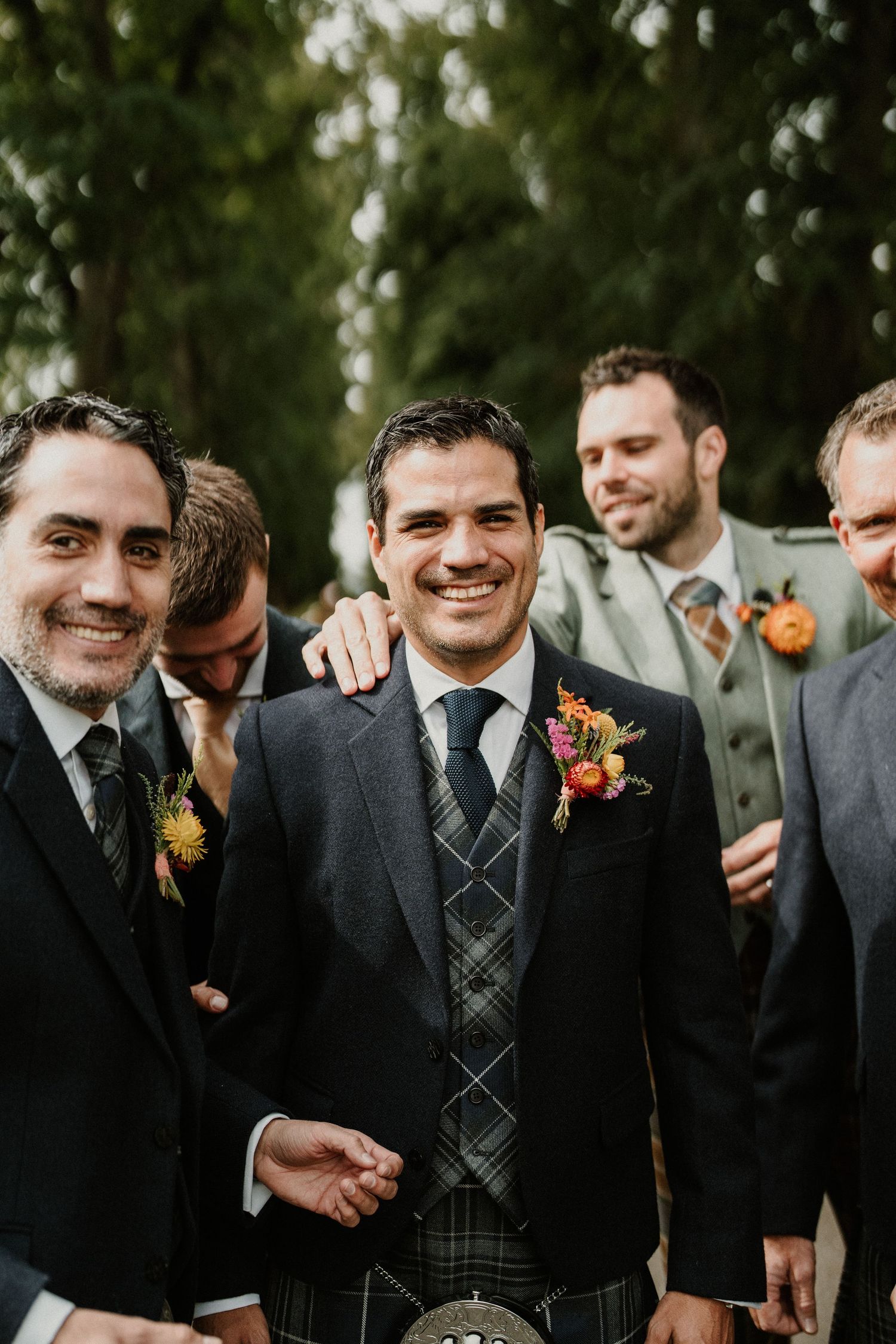 Groomsmen in dark suits with orange boutonnieres smiling outdoors.