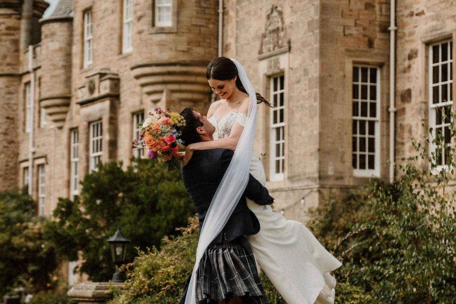 Wedding party poses together in front of a grand stone castle with beautiful landscaping.