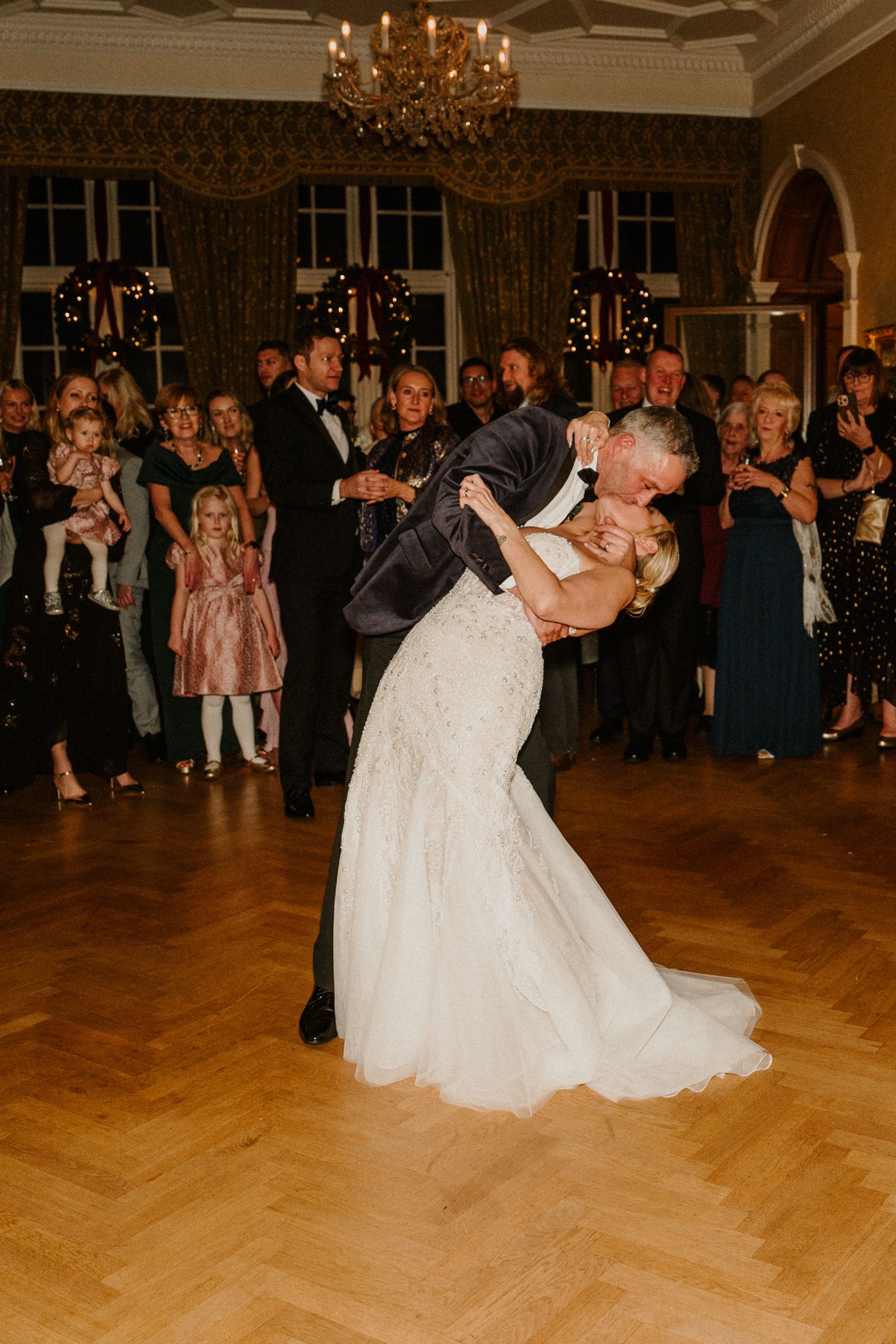 Newlyweds share a romantic dip during their first dance in an elegant ballroom with crystal chandeliers.