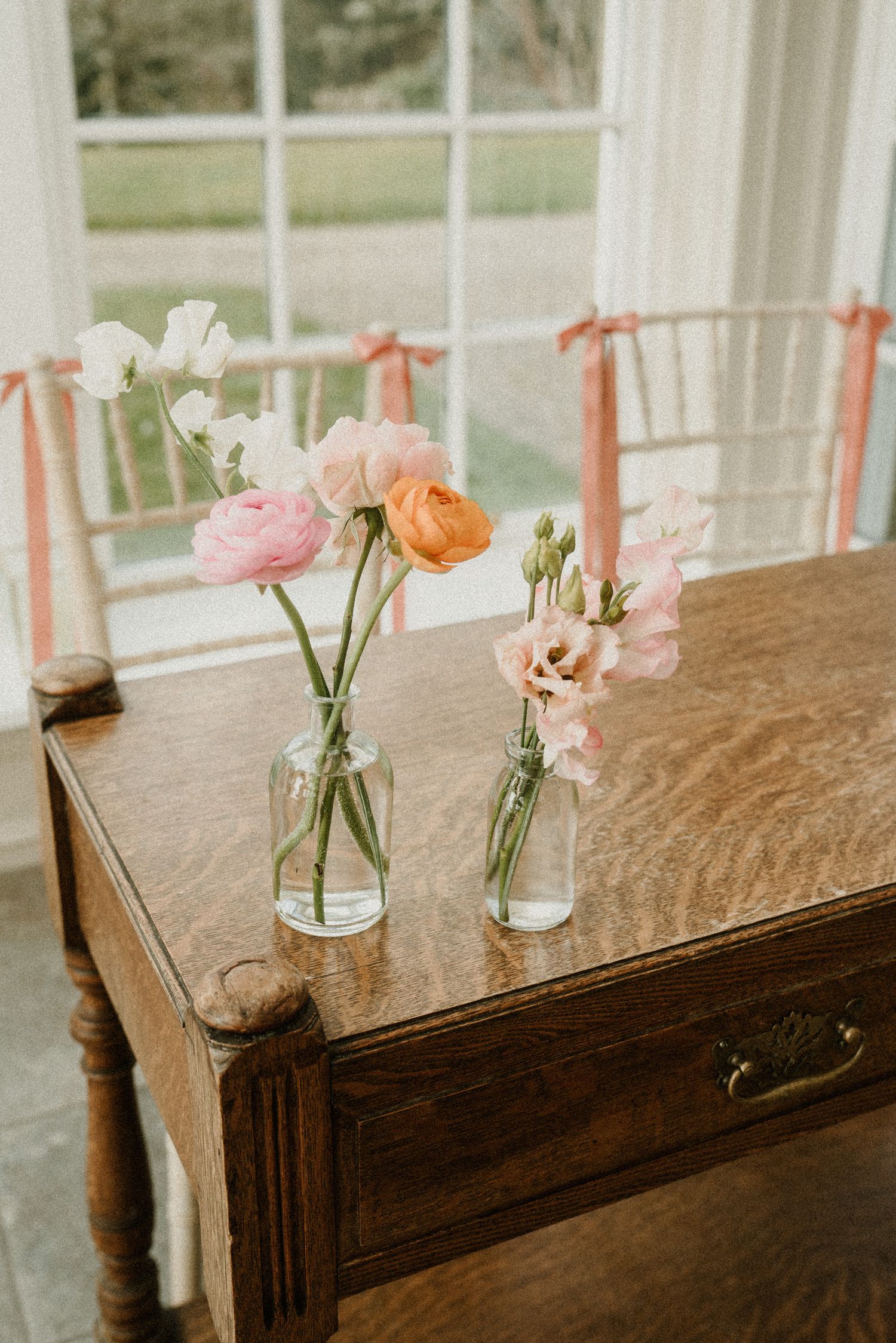 Delicate pink and peach flowers in glass vases arranged on a vintage wooden table by a window.