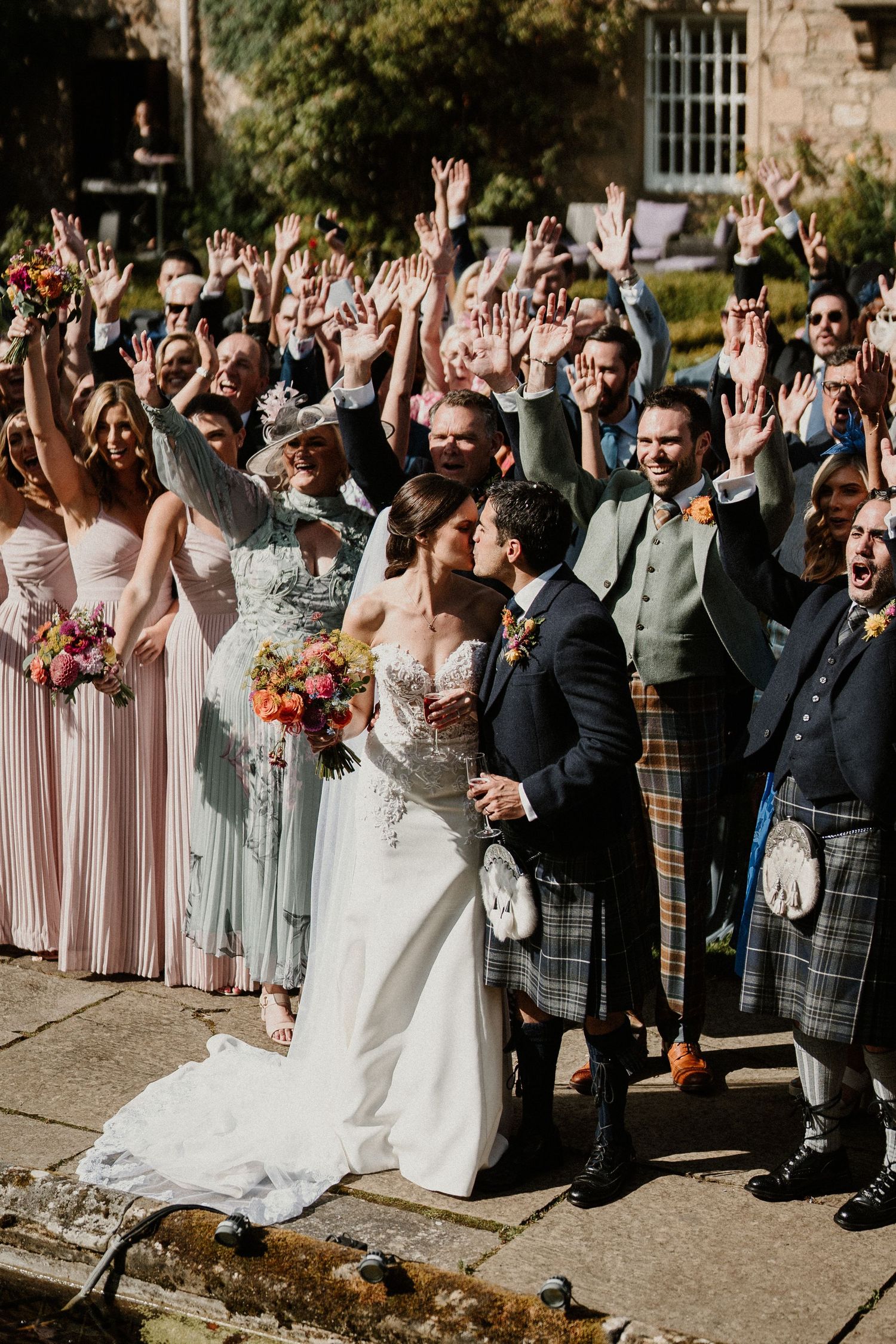 Wedding party and guests celebrating with raised hands outside historic building.