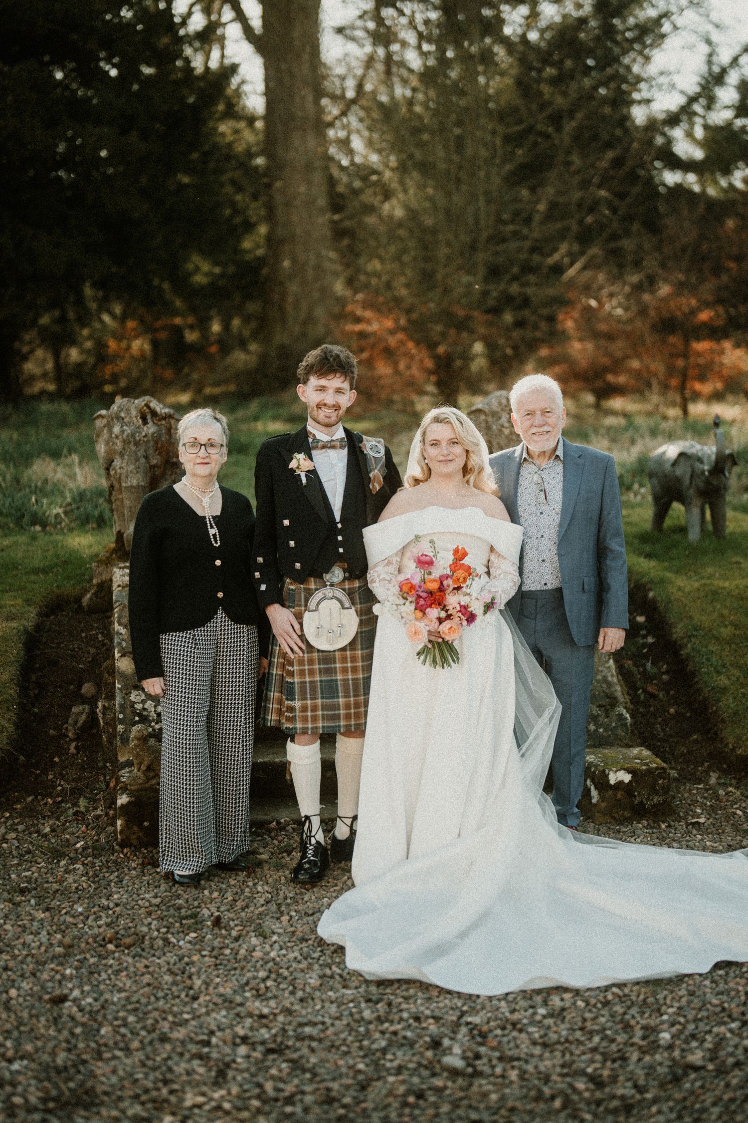Wedding party stands on gravel path with trees and autumn foliage in background.