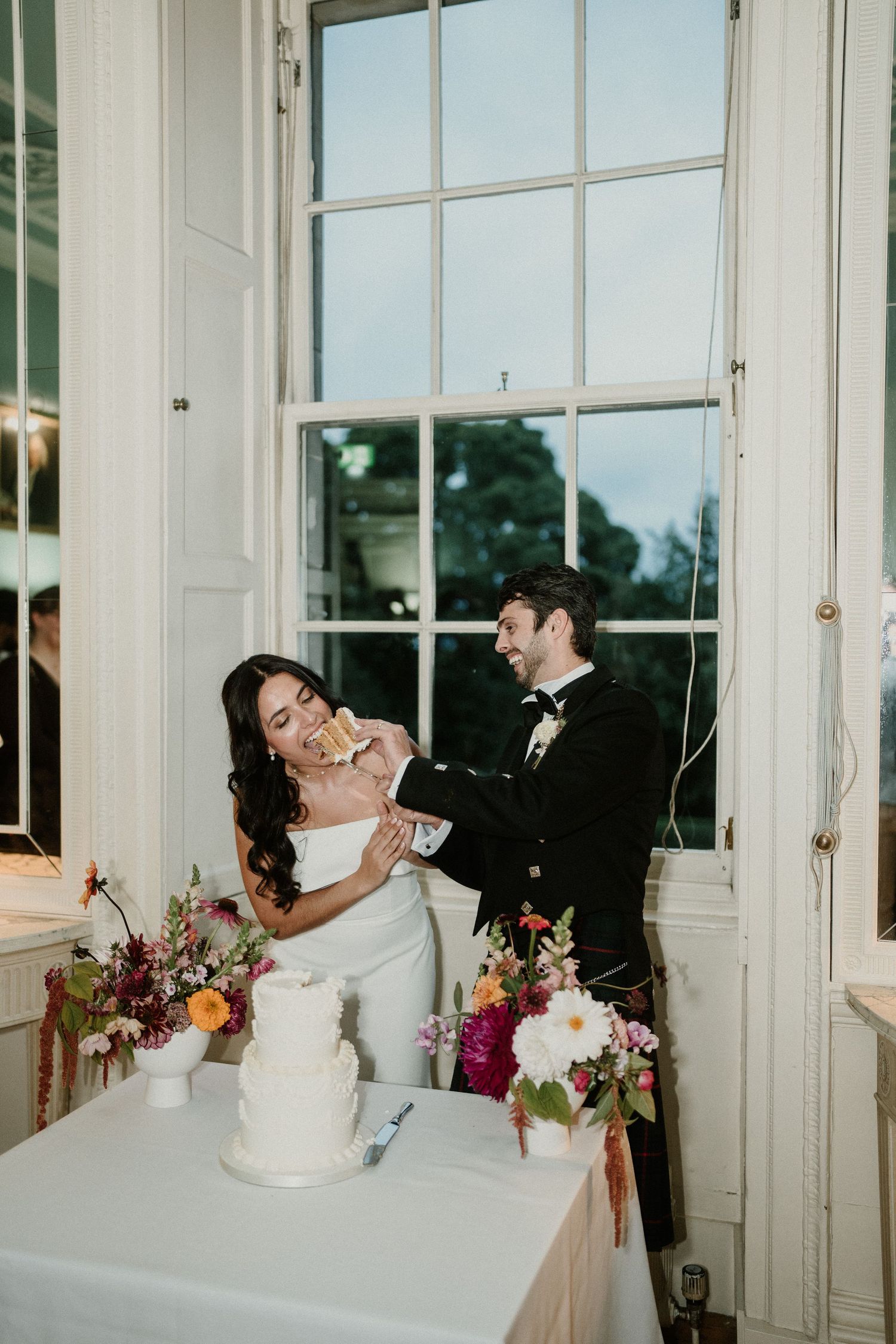 Newlyweds share a playful cake cutting moment by large windows.
