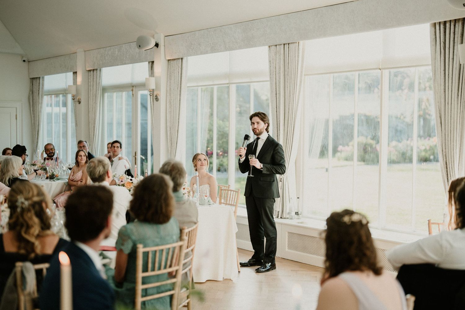 Wedding guests seated in bright sunlit reception room with large windows.