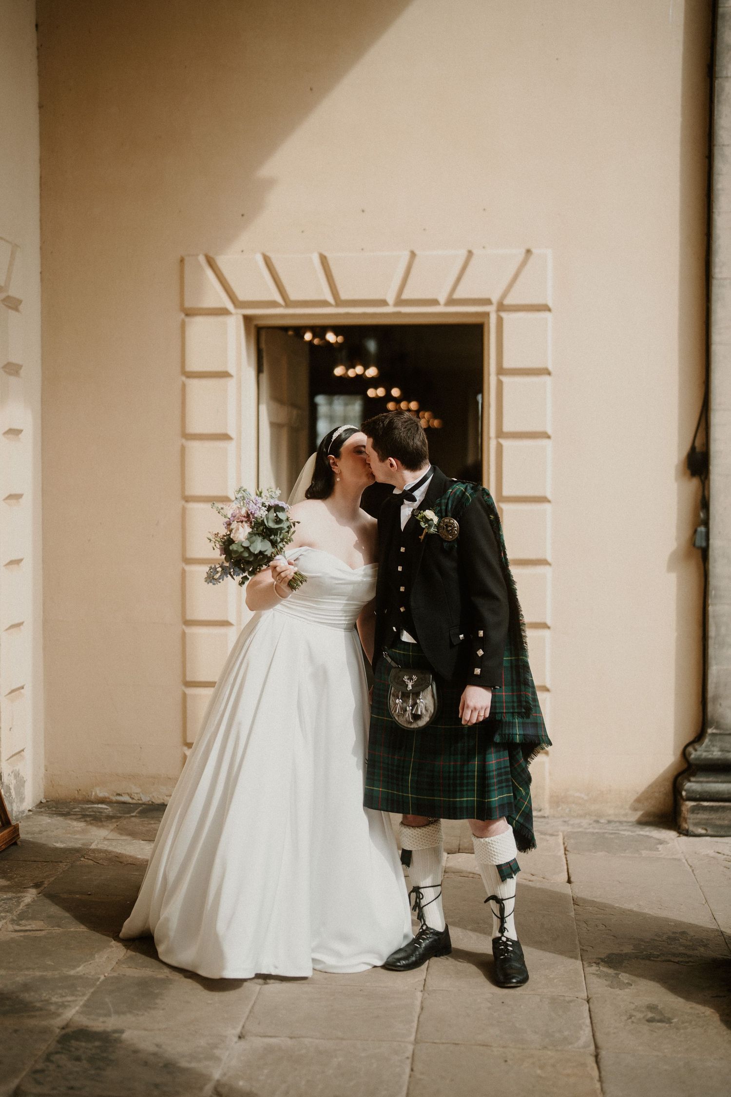 Wedding couple in traditional Scottish attire posing against a cream colored wall.
