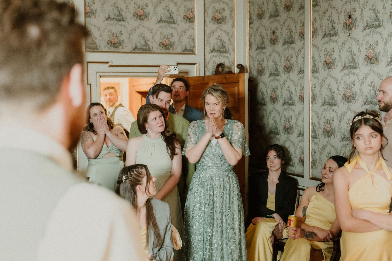 Guests in sage green and yellow dresses react emotionally during an intimate indoor wedding ceremony with vintage wallpaper.