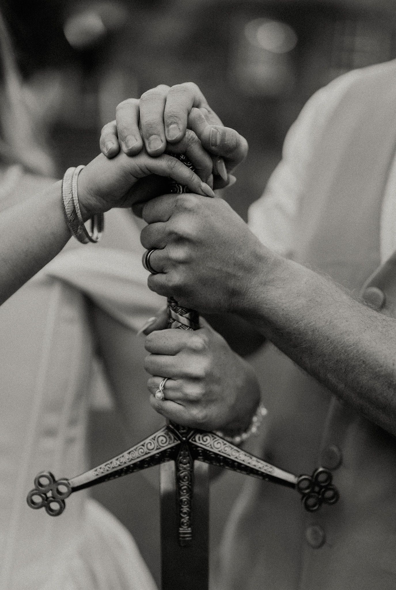 Black and white close-up of couple holding an ornate decorative sword together with wedding rings visible.