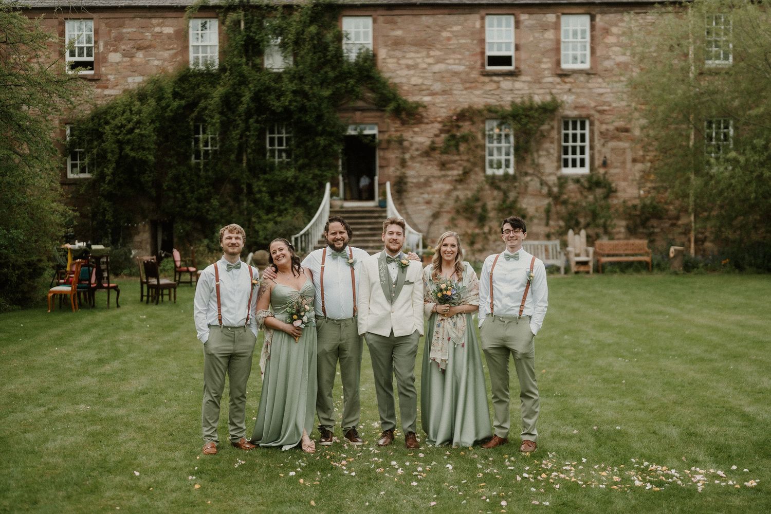 Wedding party poses outside a rustic brick manor, groomsmen in suspenders and bridesmaids in sage green gowns.