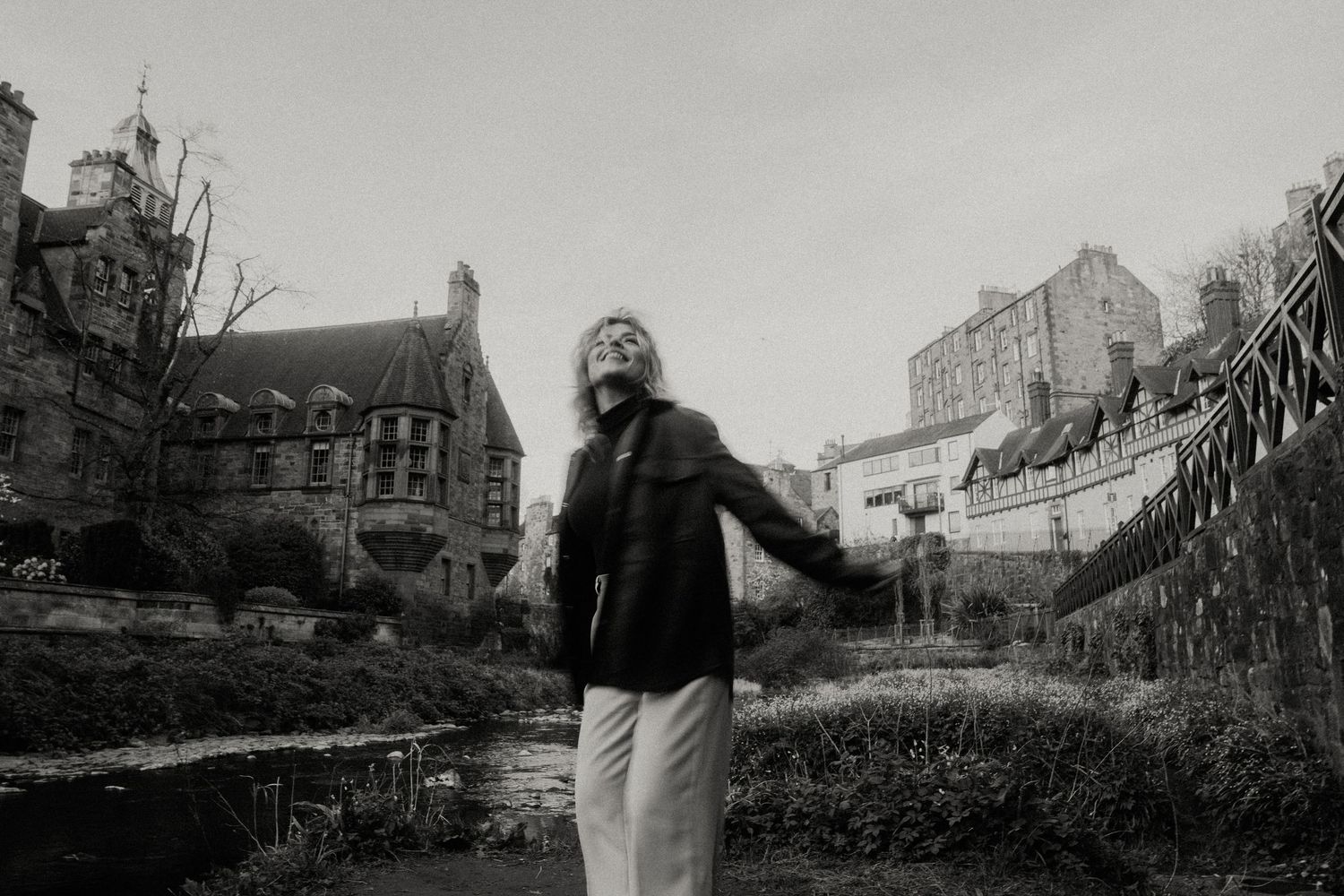 Black and white photo of a woman standing outdoors near a stream with historic stone buildings in background.