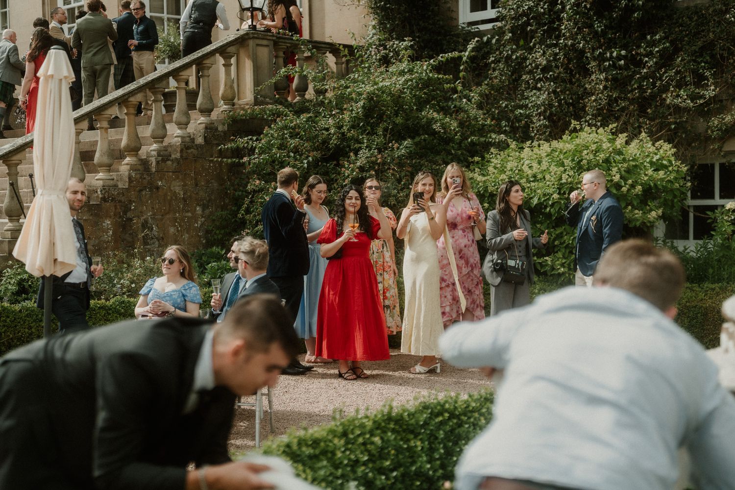 Guests in colorful formal attire gather outside a stone manor during an elegant outdoor wedding reception.